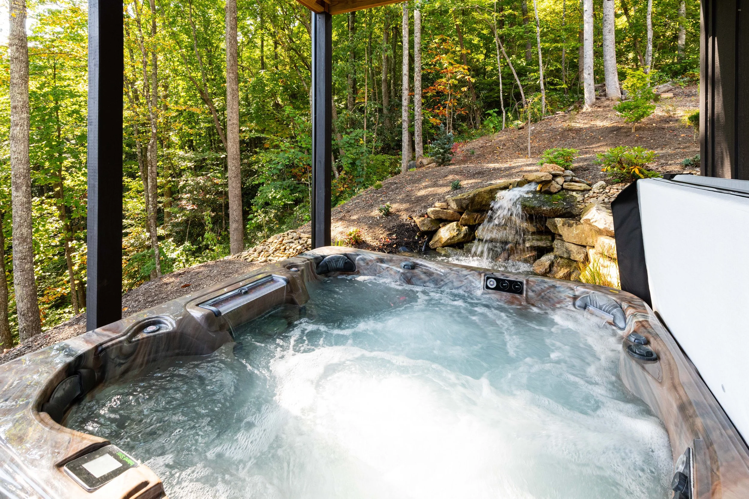 A hot tub filled with bubbling water on a patio, overlooking a wooded area with trees and a small waterfall feature.
