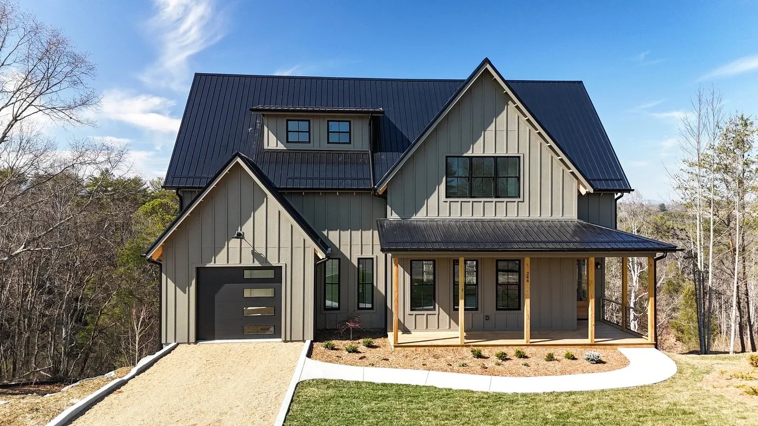 New two-story house with gray exterior, black metal roof, front porch, and a gravel driveway, surrounded by trees and a lawn.