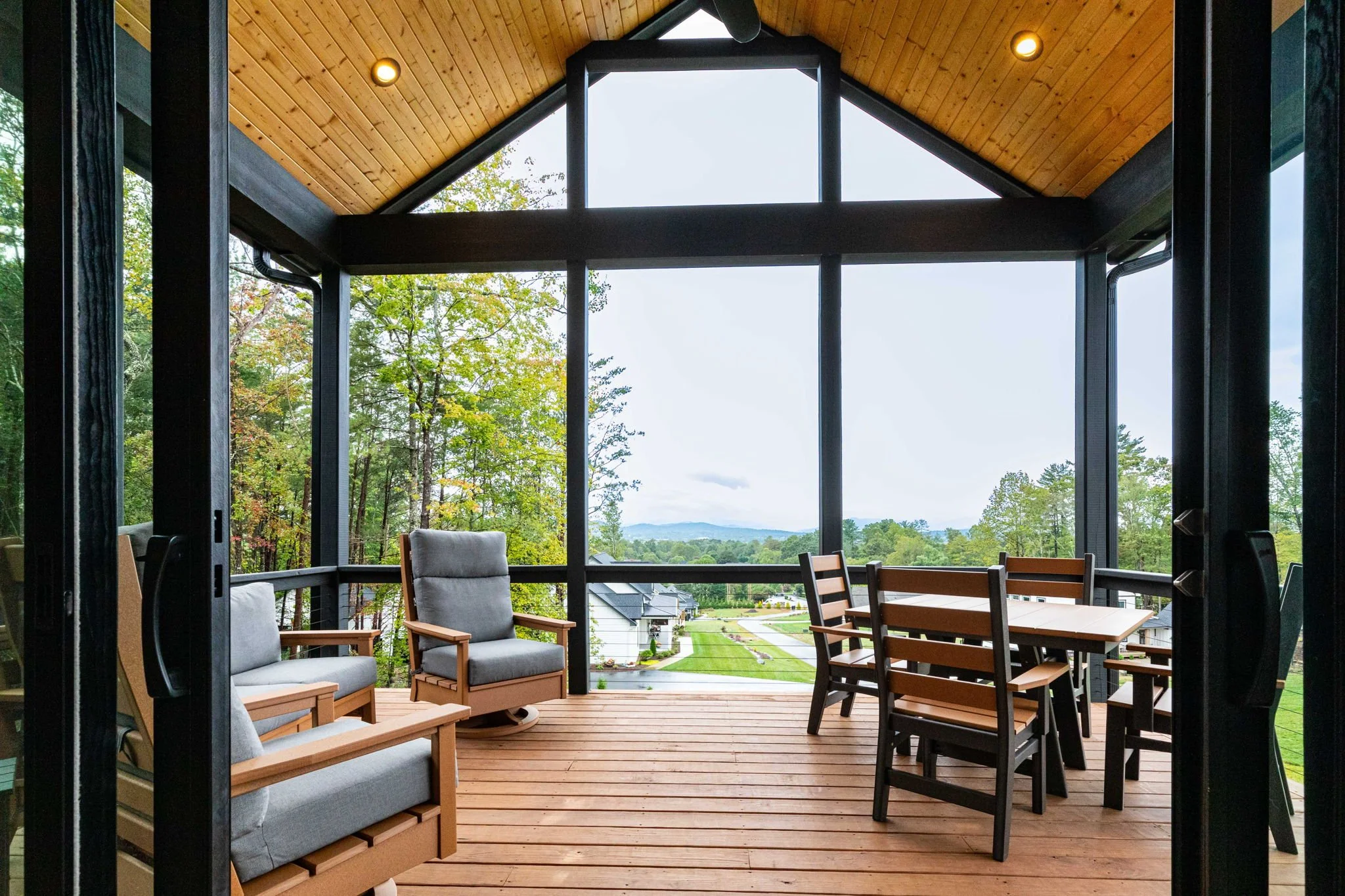 Covered wooden porch with outdoor furniture, including cushioned chairs and a table with chairs, overlooking a suburban neighborhood and green landscape with trees and hills.