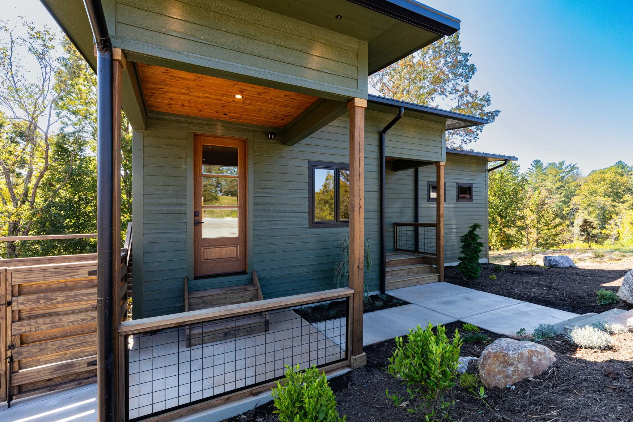 A modern house with a green exterior, wooden accents, small porch, and steps leading up to the front door, surrounded by trees and landscaping.