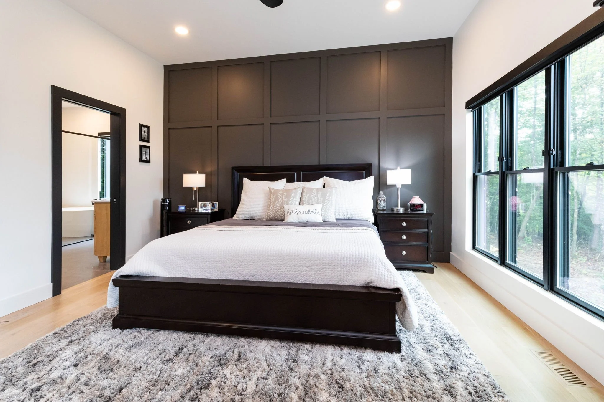 A bedroom with a dark wooden bed frame, white bedding, and multiple pillows, flanked by matching nightstands with lamps. There is a large window on the right side, and a door on the left leading to another room, possibly a bathroom.