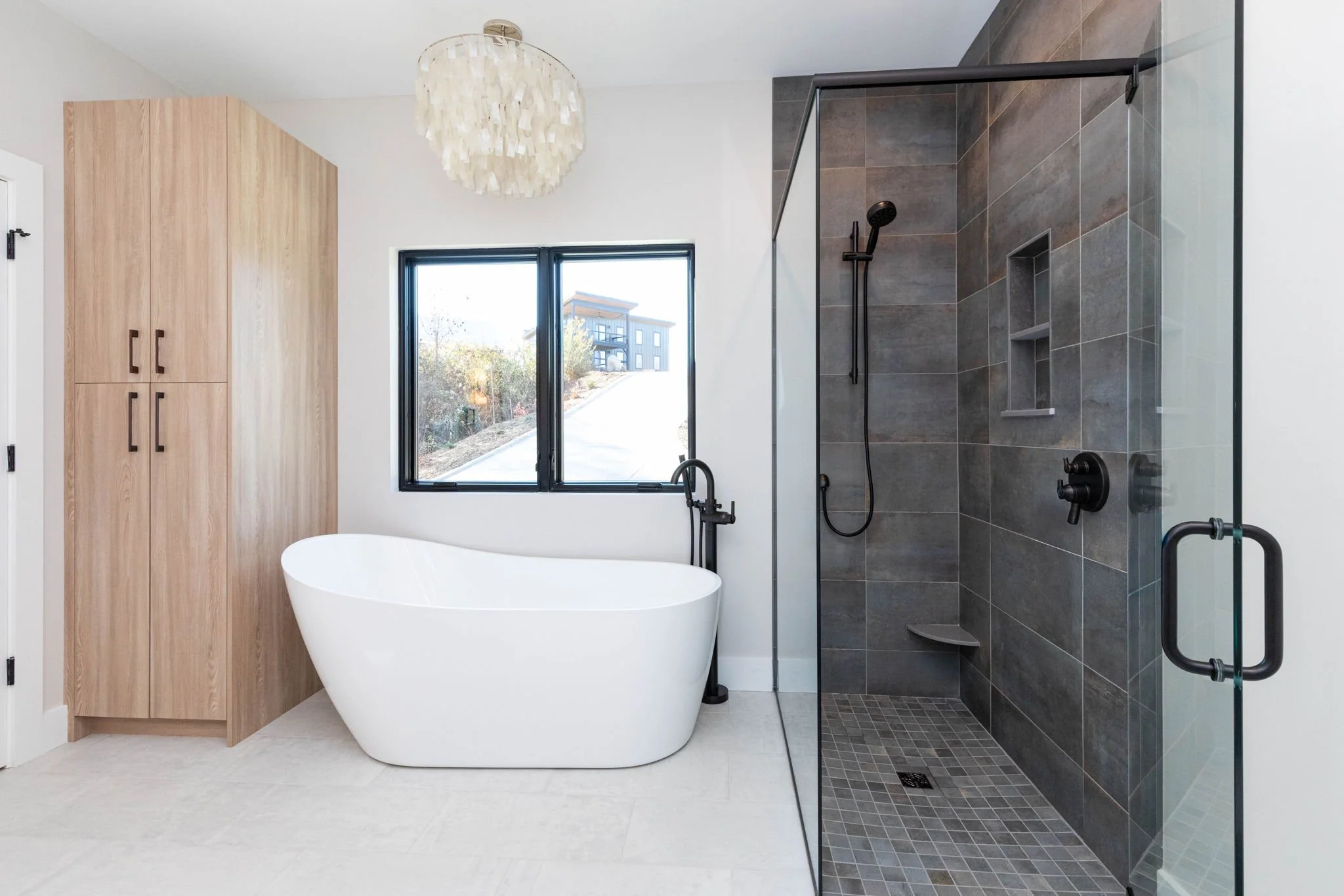 Modern bathroom featuring a freestanding bathtub, a walk-in shower with dark gray tiles and glass door, a black window, a light wooden cabinet, and a white chandelier.