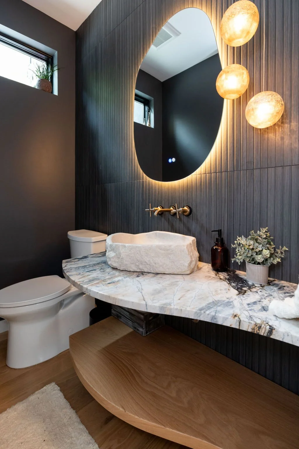 Modern bathroom with dark textured wall, large oval mirror with backlight, three spherical pendant lights, stone vessel sink on marble countertop, white toilet, small potted plant, and soap dispenser.