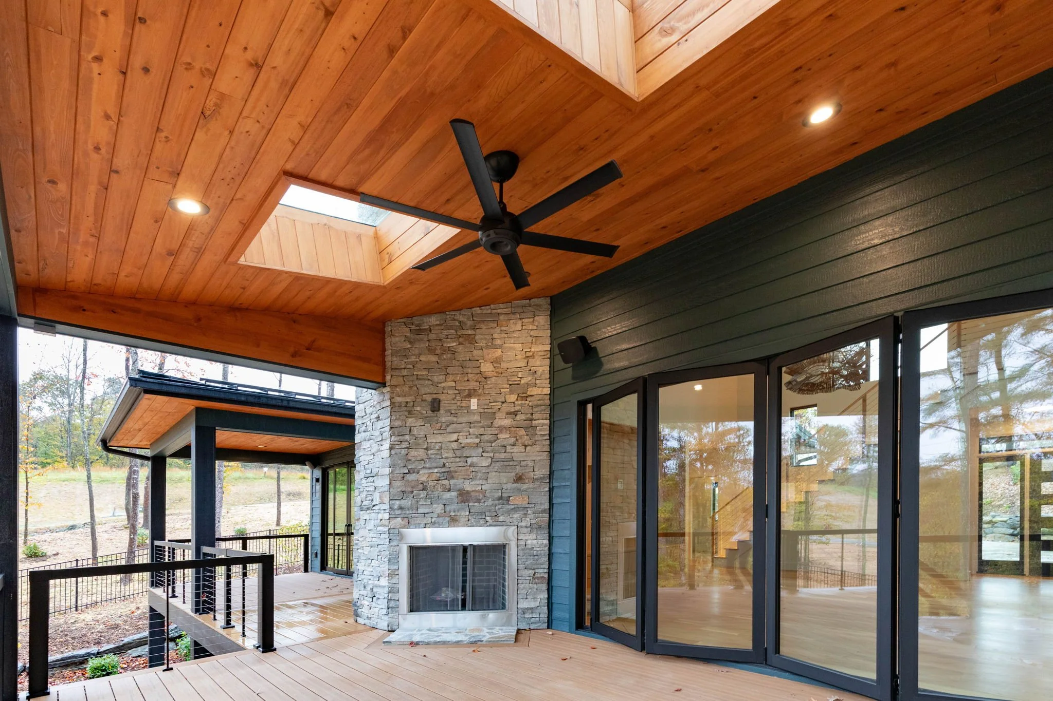 Outdoor view of a modern house with a wooden ceiling, ceiling fan, skylight, stone fireplace, and large glass sliding doors.