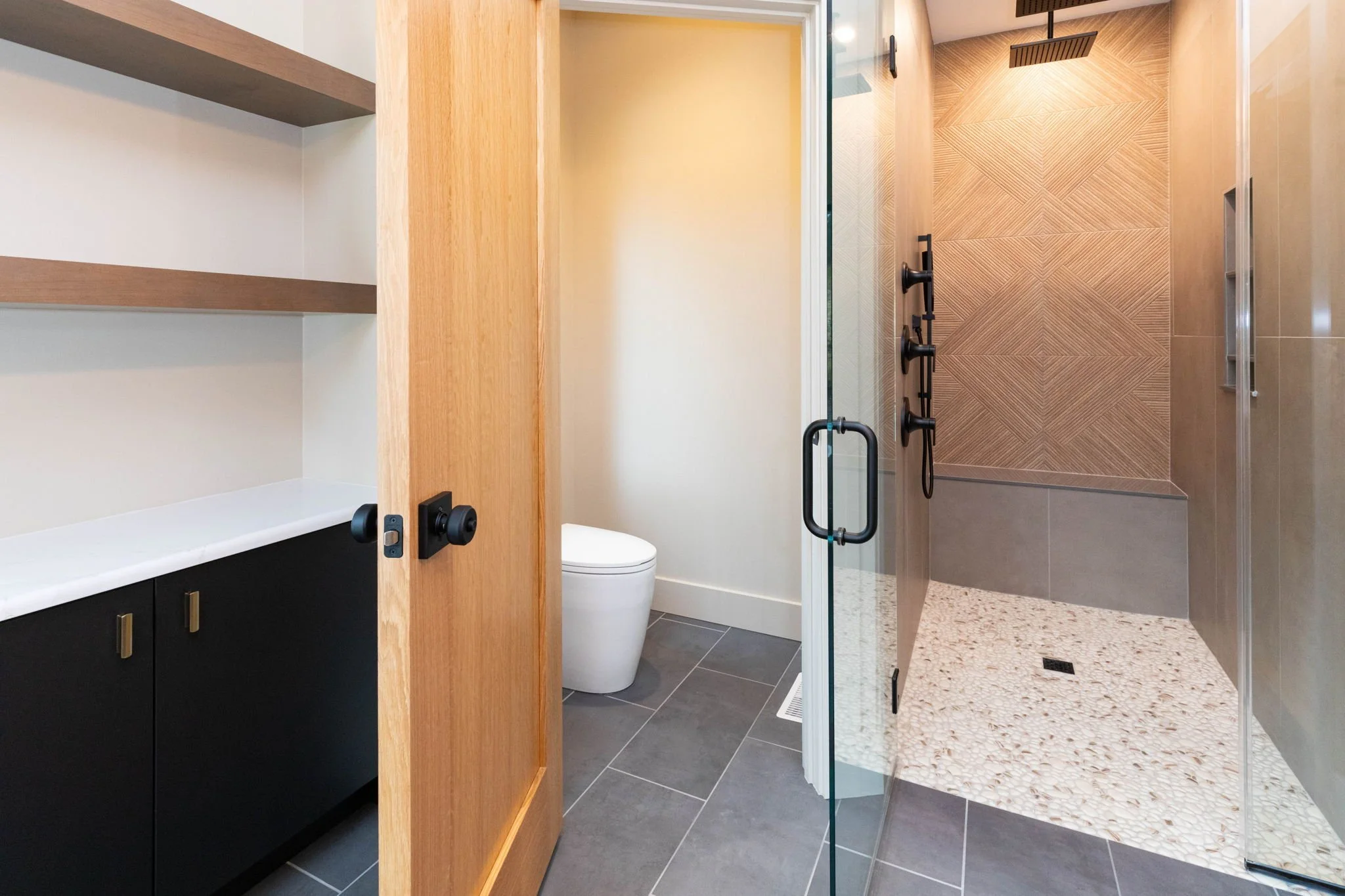 Modern bathroom with a glass walk-in shower with textured wall tiles, a rain showerhead, and a black fixture. A black and white cabinet with open shelves and a countertop, and a white toilet are visible.