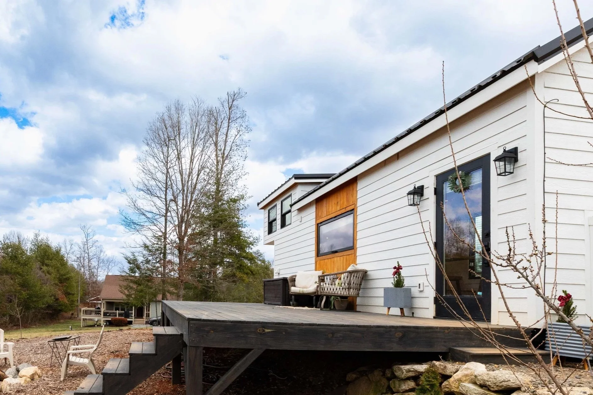 Exterior view of a modern two-story house with a wooden and white siding deck, outdoor furniture, and a tree with budding branches. The sky is cloudy and other houses are visible in the background.