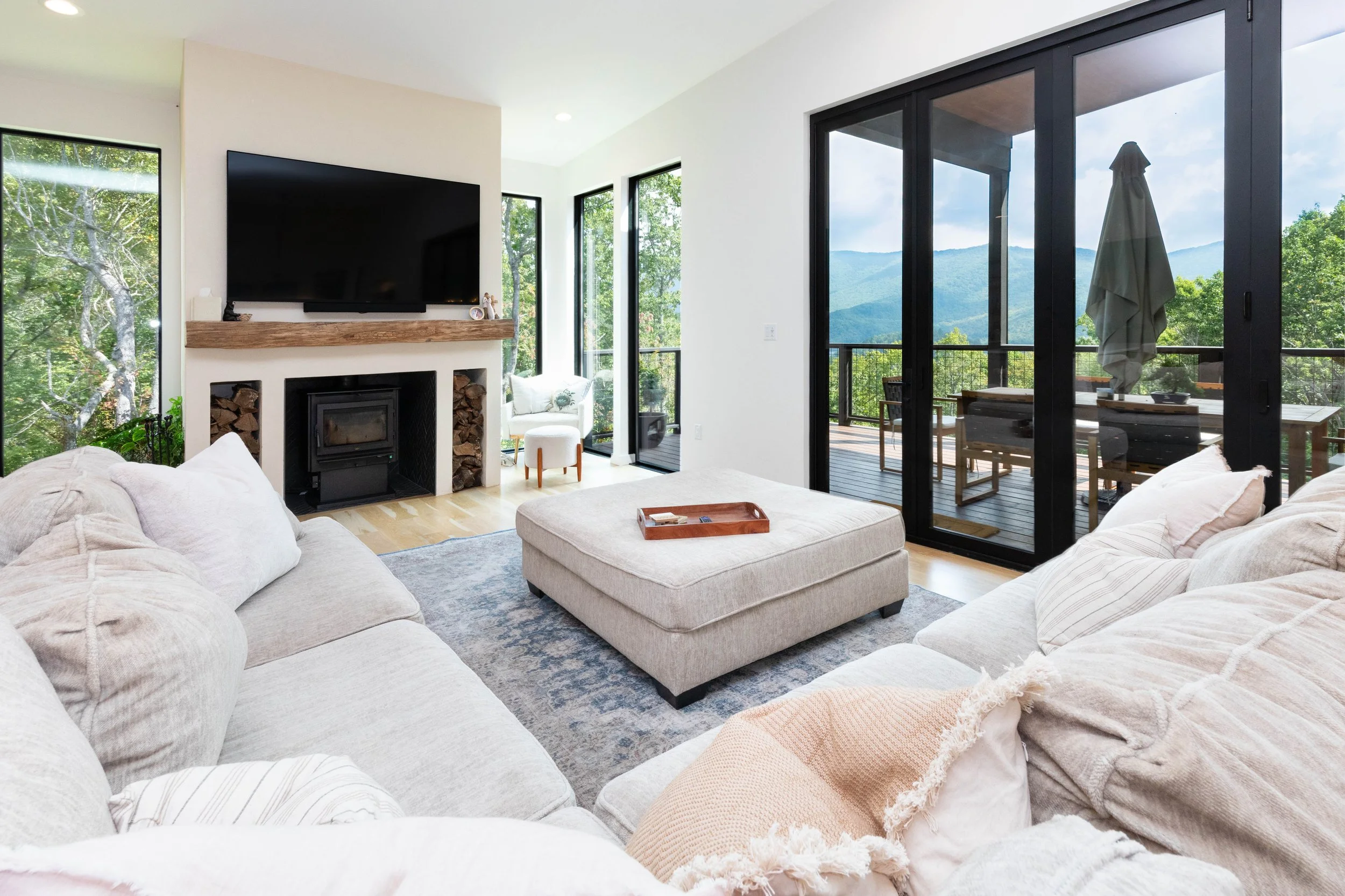 Bright living room with large sliding glass doors opening to a deck with mountain views, a beige sectional sofa with pillows, a rectangular ottoman, a fireplace with a mounted TV above, and windows showing trees outside.