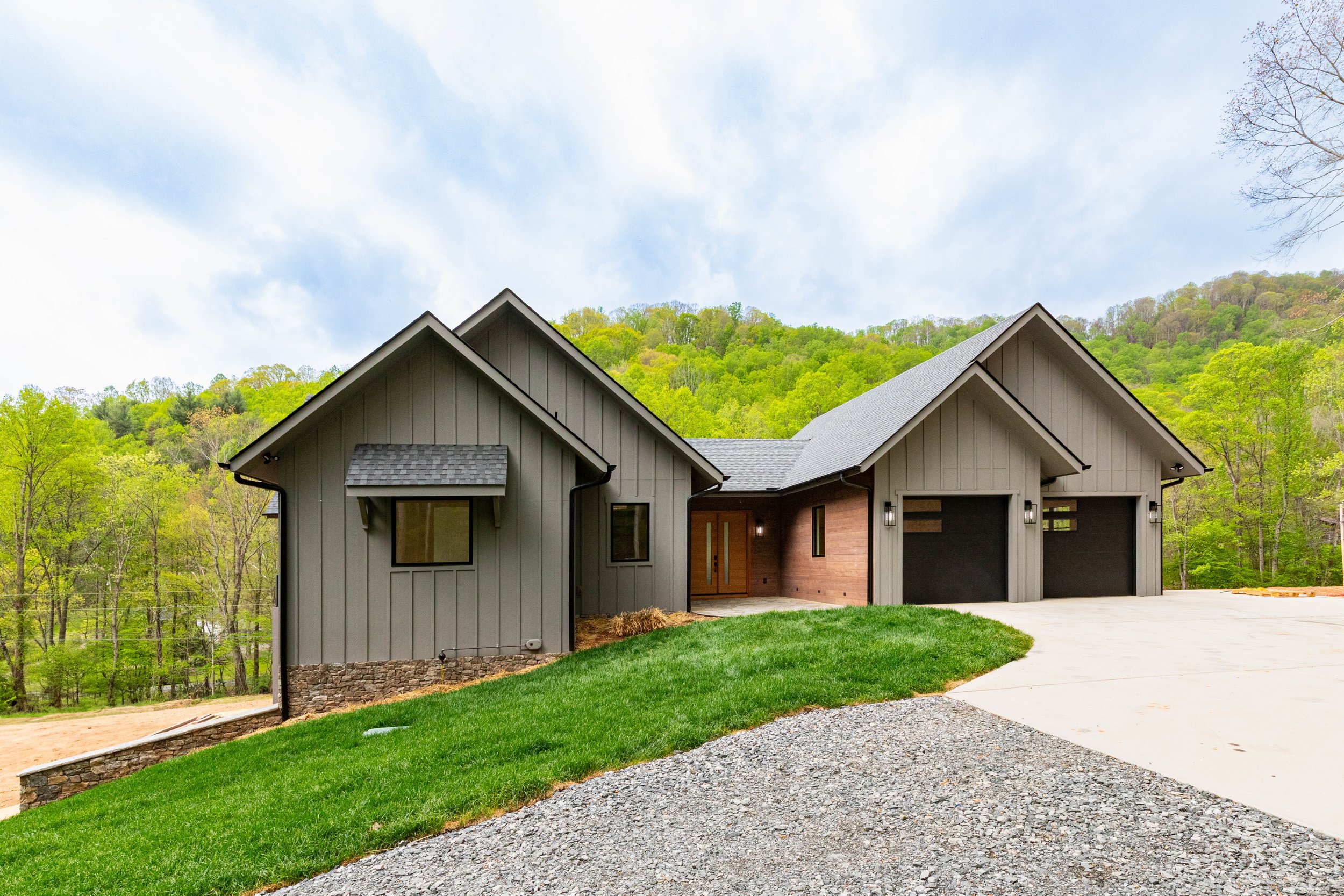Modern house with gray vertical siding, stone accents, and a double garage, surrounded by greenery and trees, with a driveway and a grassy yard.