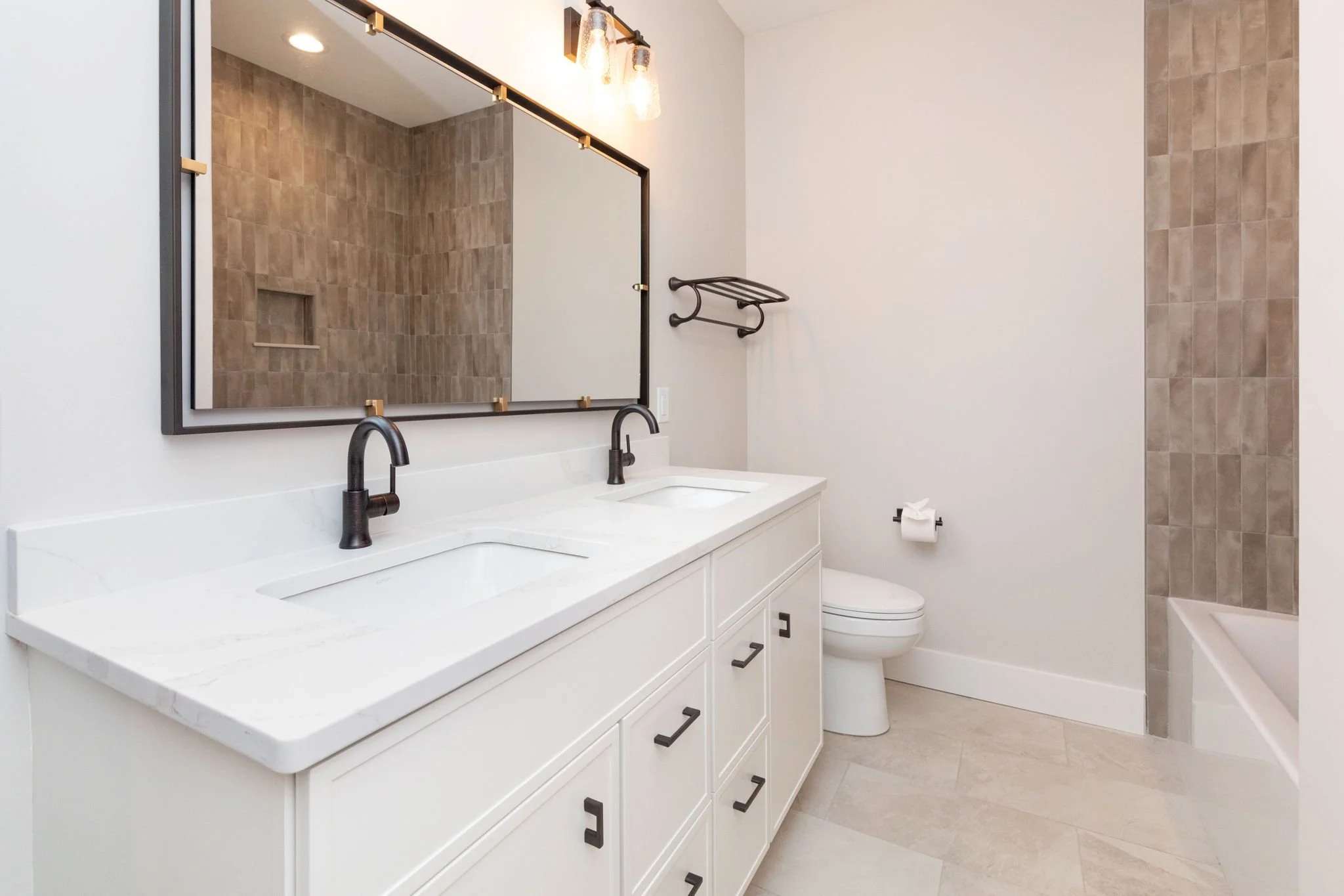 Modern bathroom with double vanity, black faucets, large mirror, beige tile shower, white walls, and a toilet.