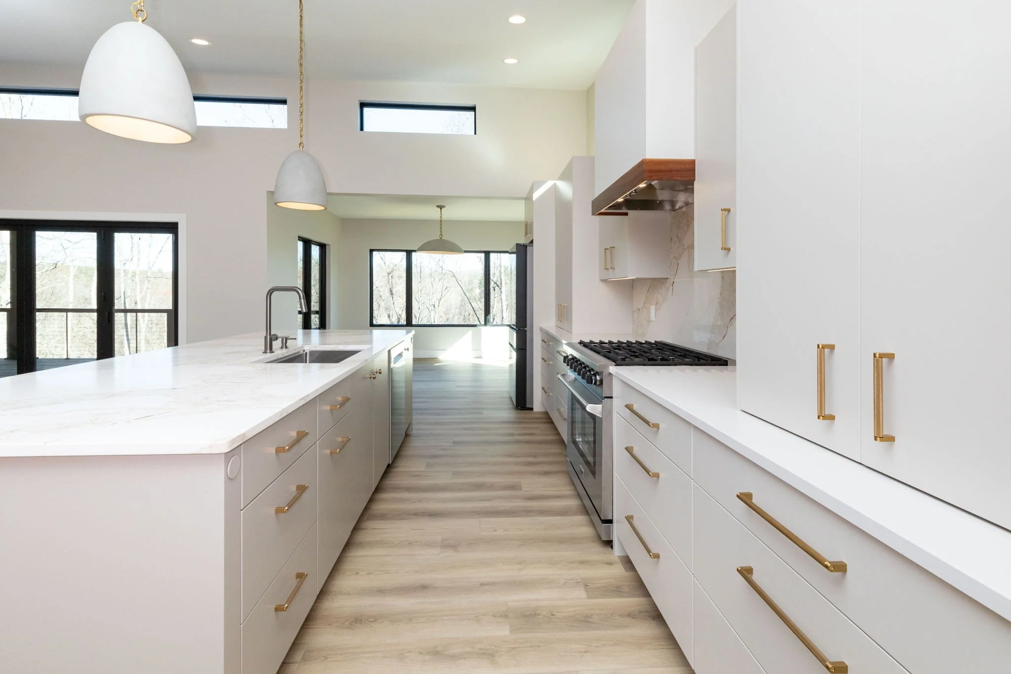 Modern minimalist kitchen with white cabinets, marble countertops, wood accents, and large windows in the background.