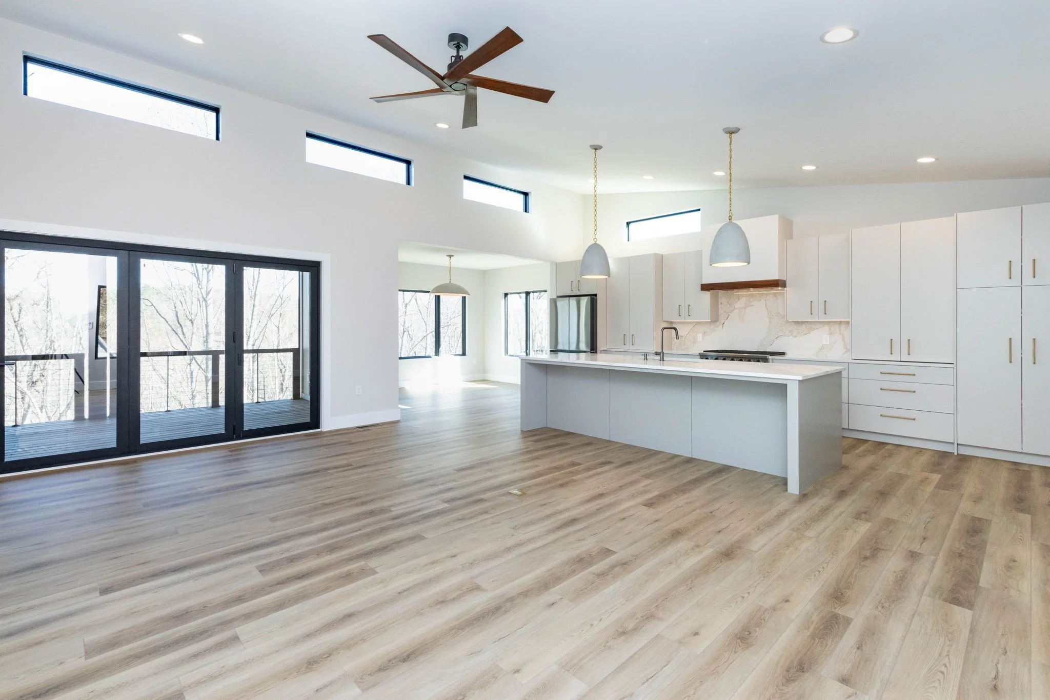 Open-concept modern kitchen and living area with light wood flooring, white cabinetry, large sliding glass doors leading to a balcony, and high horizontal windows.