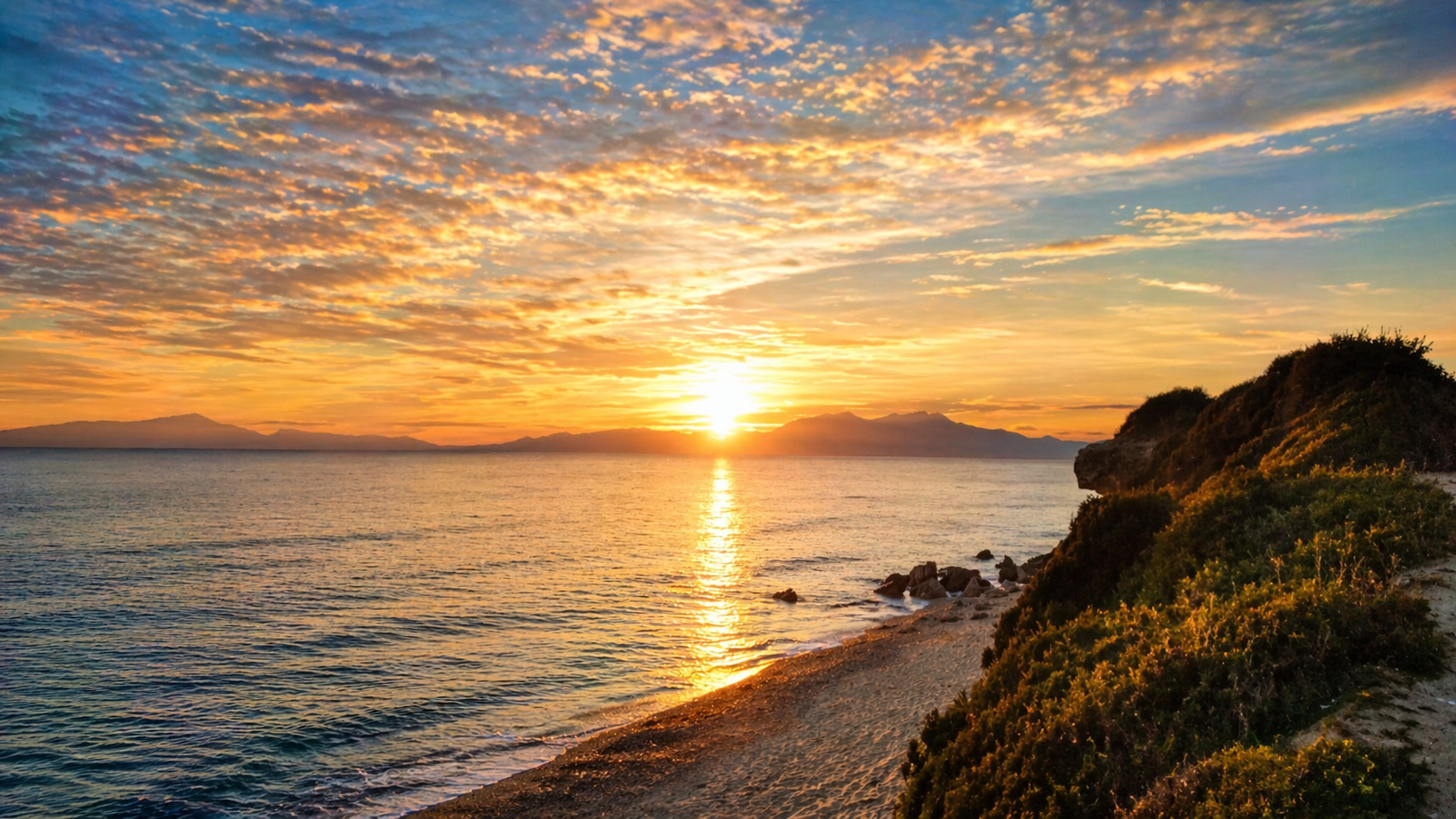 Sunset over the ocean with cloudy sky, rocky shoreline, and green bushes in the foreground.