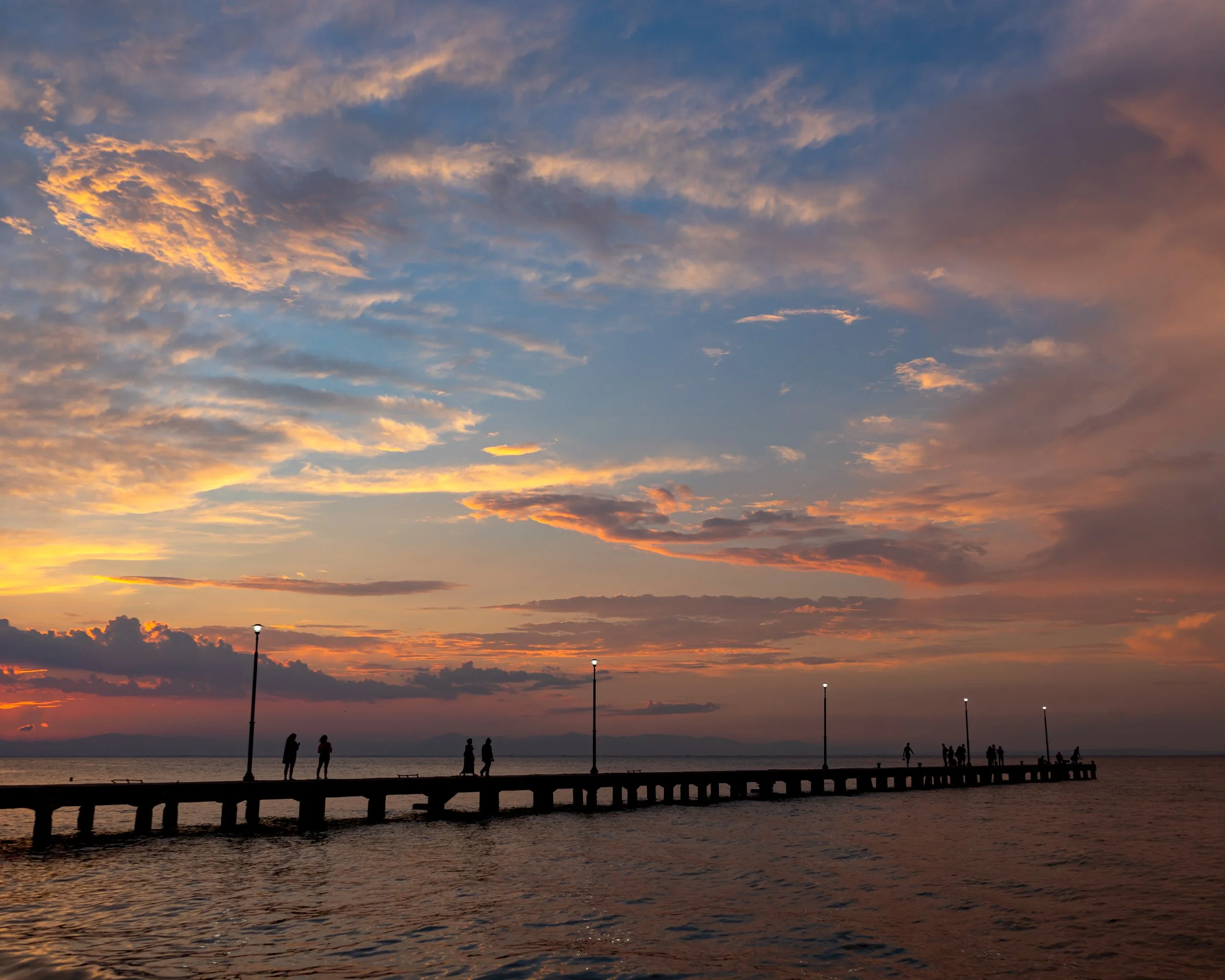 Sunset over the ocean with colorful clouds in the sky. Silhouettes of people walking on a pier and some on the beach at dusk at Nees Epivates, Pereia, Thessaloniki