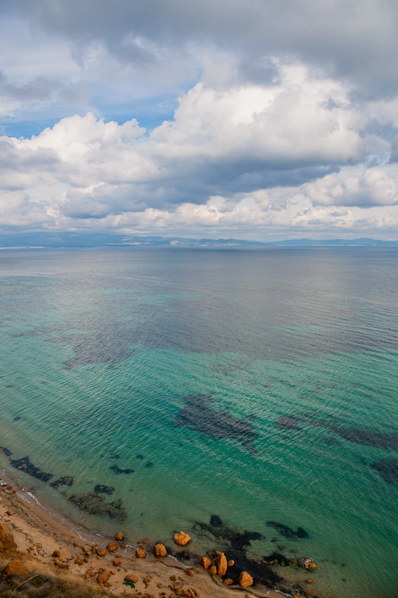 A view of a coastline with sandy beach, scattered rocks, and clear green water under a cloudy sky.