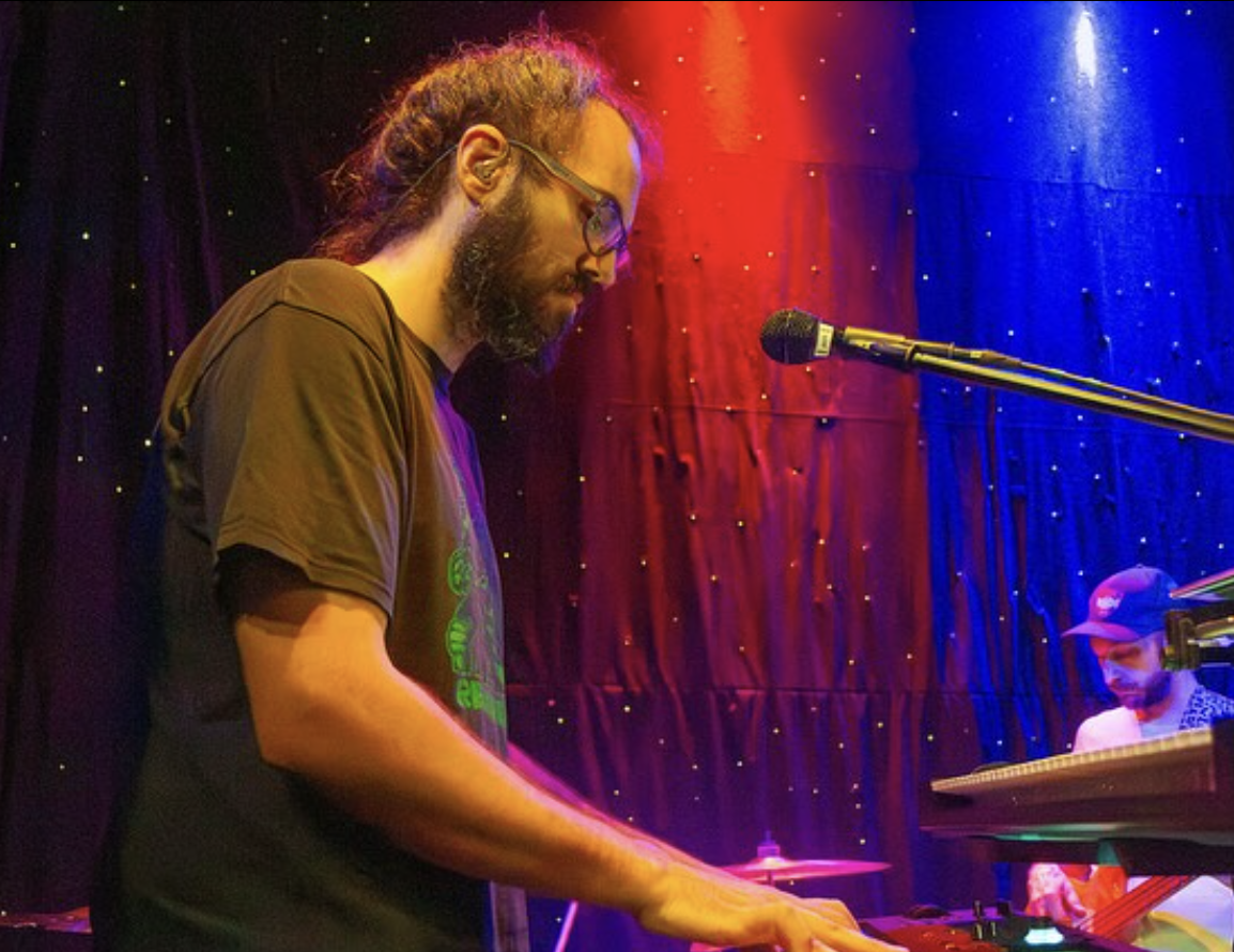 Man with hair pulled back wearing a black t-shirt is playing the keyboard with a mic in front of him