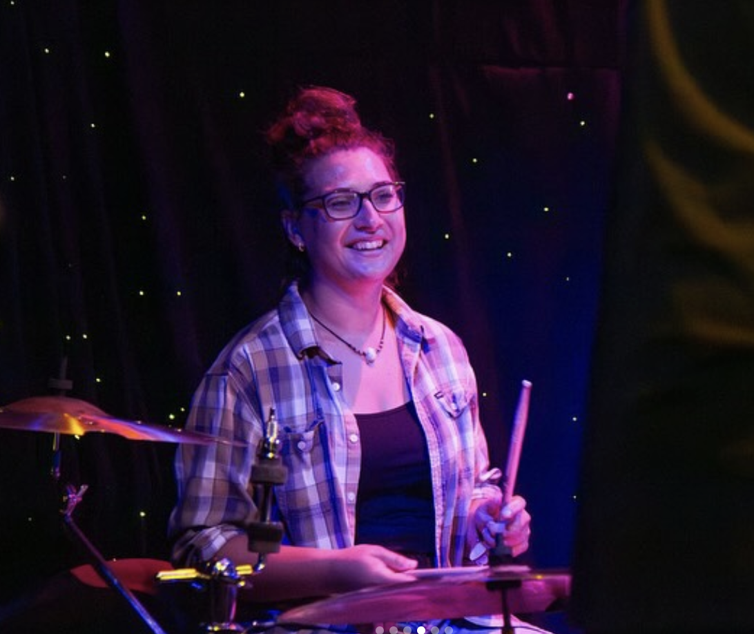A woman is sitting at a drumset holding drum sticks and smiling wearing a flannel