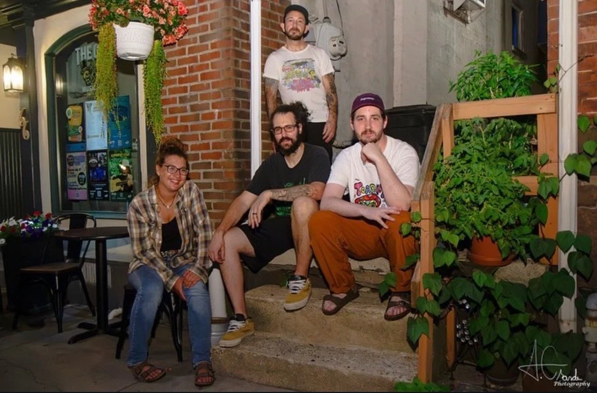 Three men and one woman sit outstairs on a stoop looking at the camera
