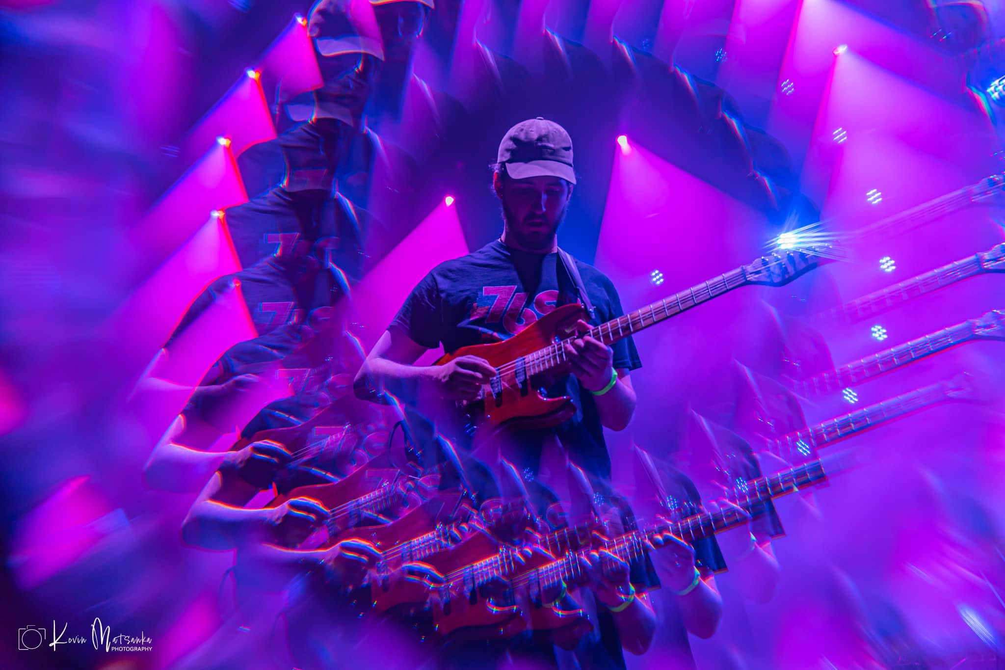 A man wearing a baseball cap playing guitar in a purple and pink swirl of lights