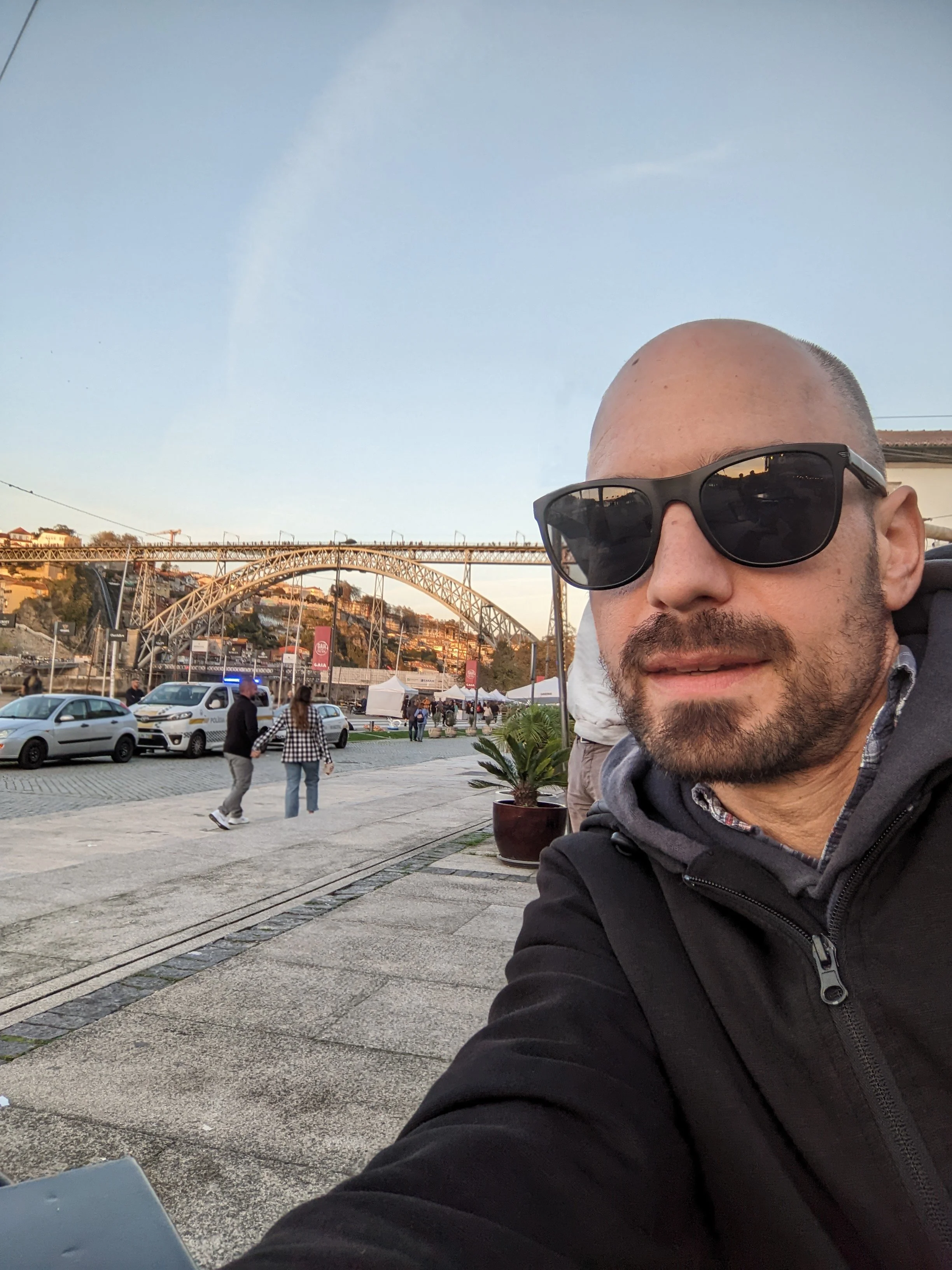 A man in sunglasses and a black jacket taking a selfie outdoors in Porto, Portugal with a cityscape, bridge, and blue sky in the background.