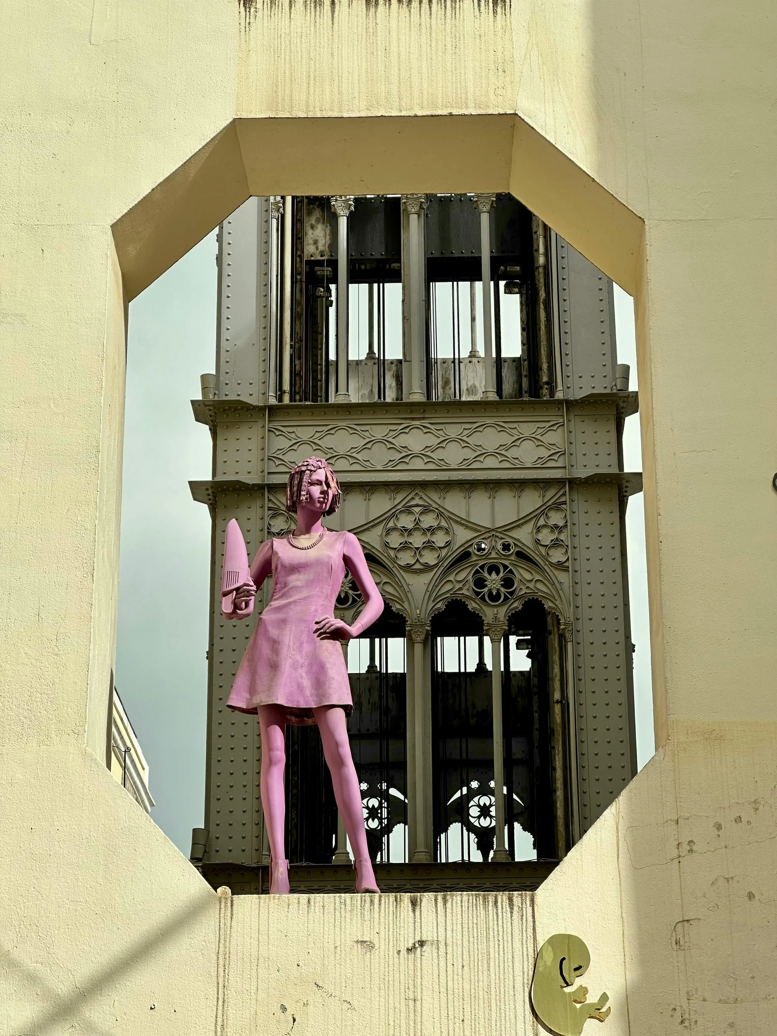 A pink statue of a woman holding a book, standing in a large, open window frame with architectural details and an elevator shaft behind her.