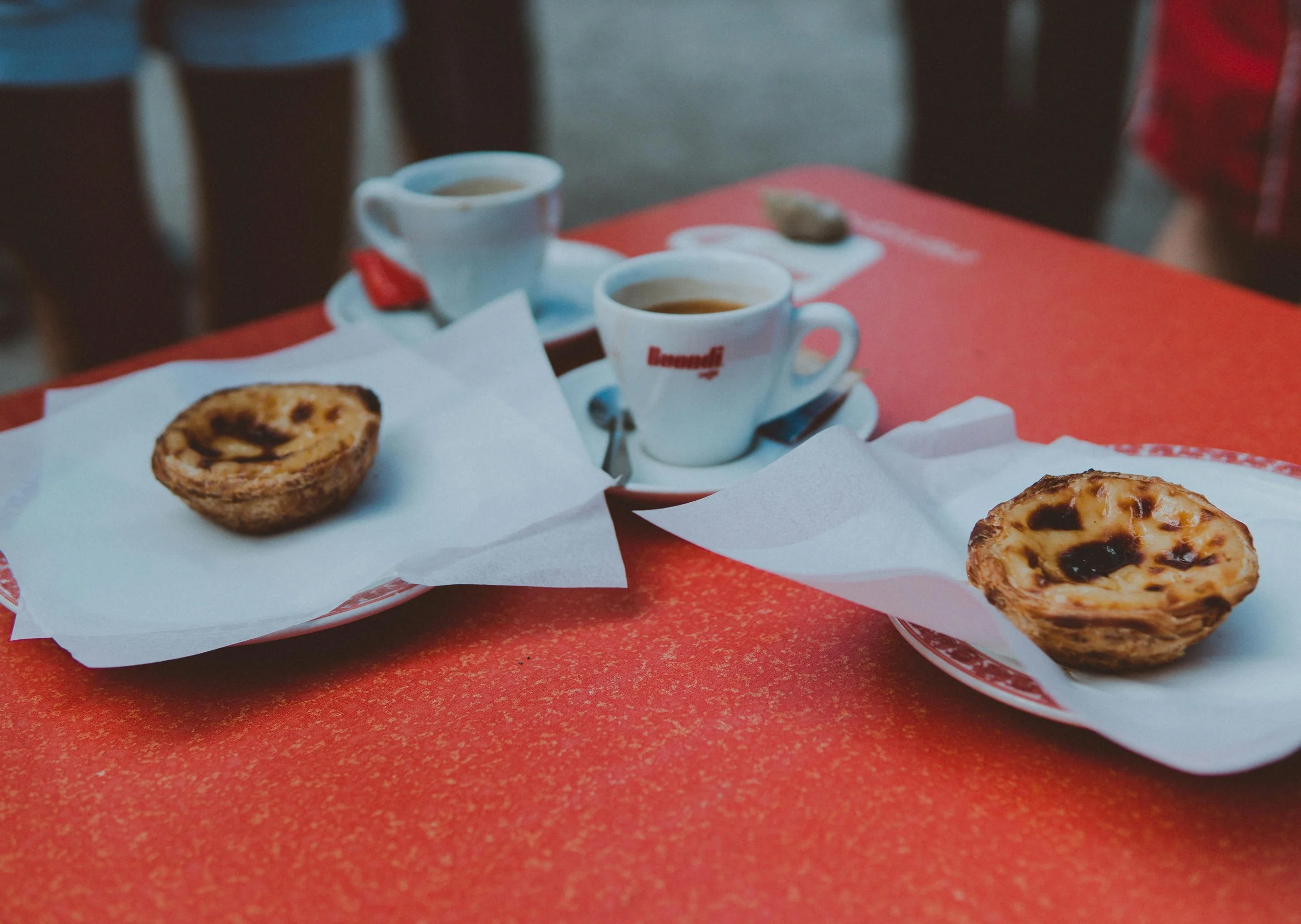 Two Portuguese espressos in Buondi brand cups on a red café table, along with two pasteis de nata.