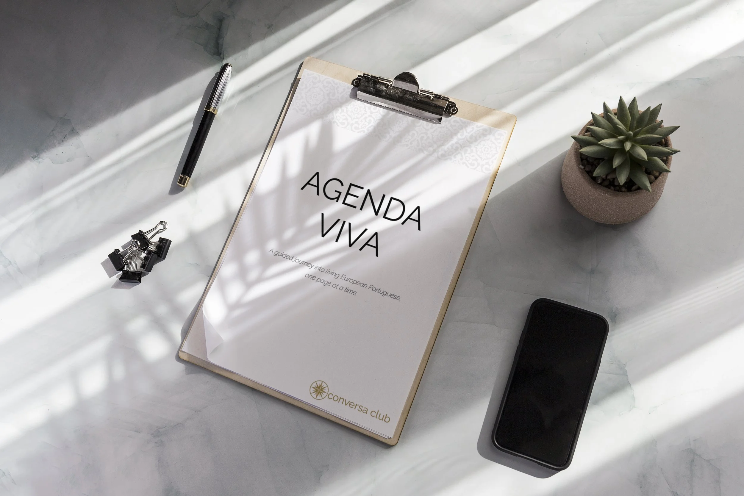 A clipboard with a paper titled 'Agenda Viva' on a white marble surface, accompanied by a pen, a black smartphone, three black binder clips, and a potted succulent plant, with sunlight casting shadows across the items.