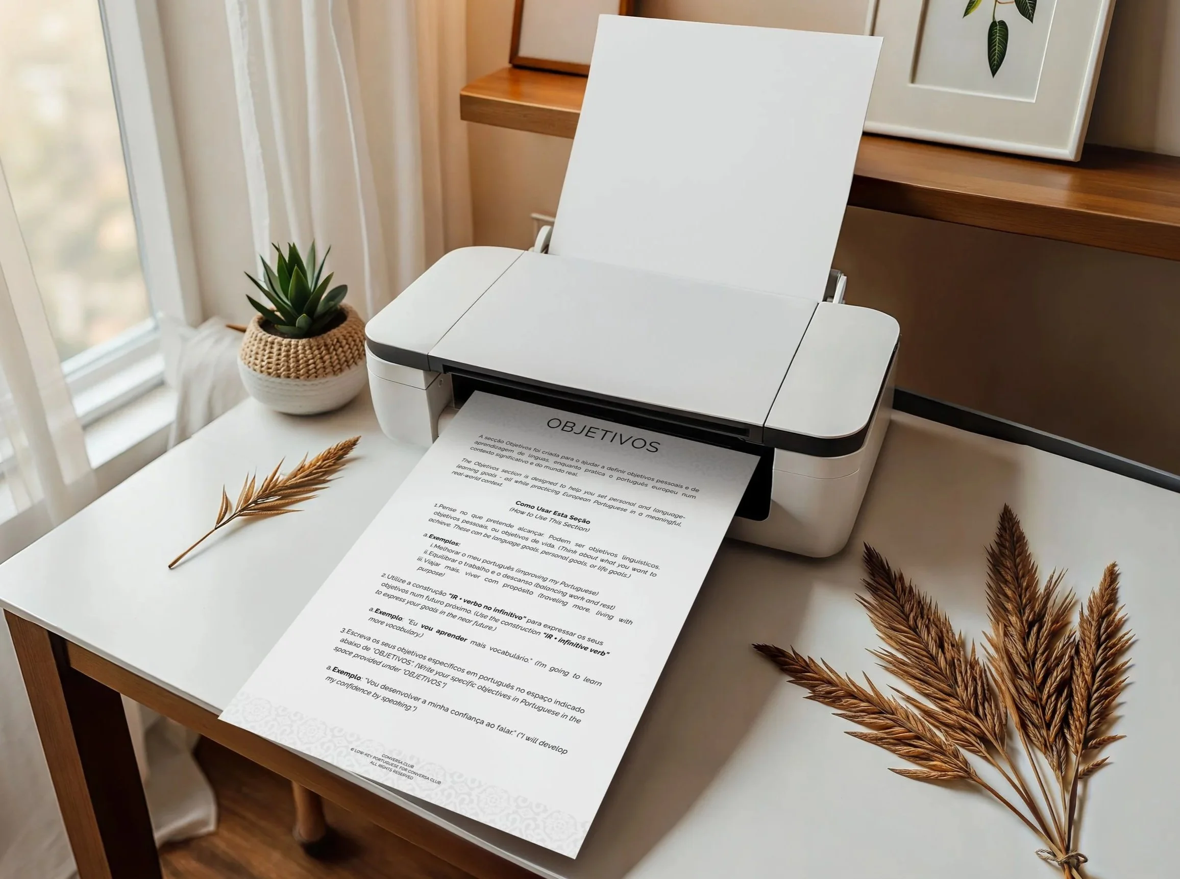 A white desktop printer on a table with a printed document titled 'OBJETIVOS' being fed through, surrounded by decorative dried wheat stalks, a small potted plant, and a framed botanical print, with a window and curtains in the background.