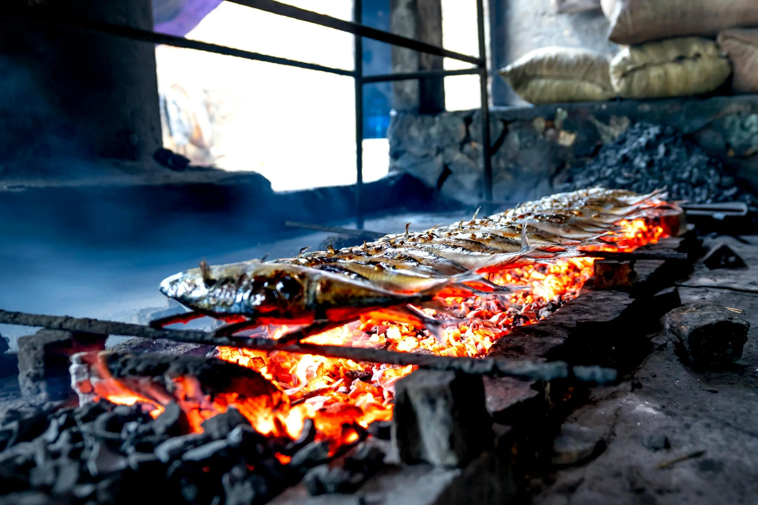Fish being grilled on an open flame with glowing embers, inside a rustic outdoor cooking area.