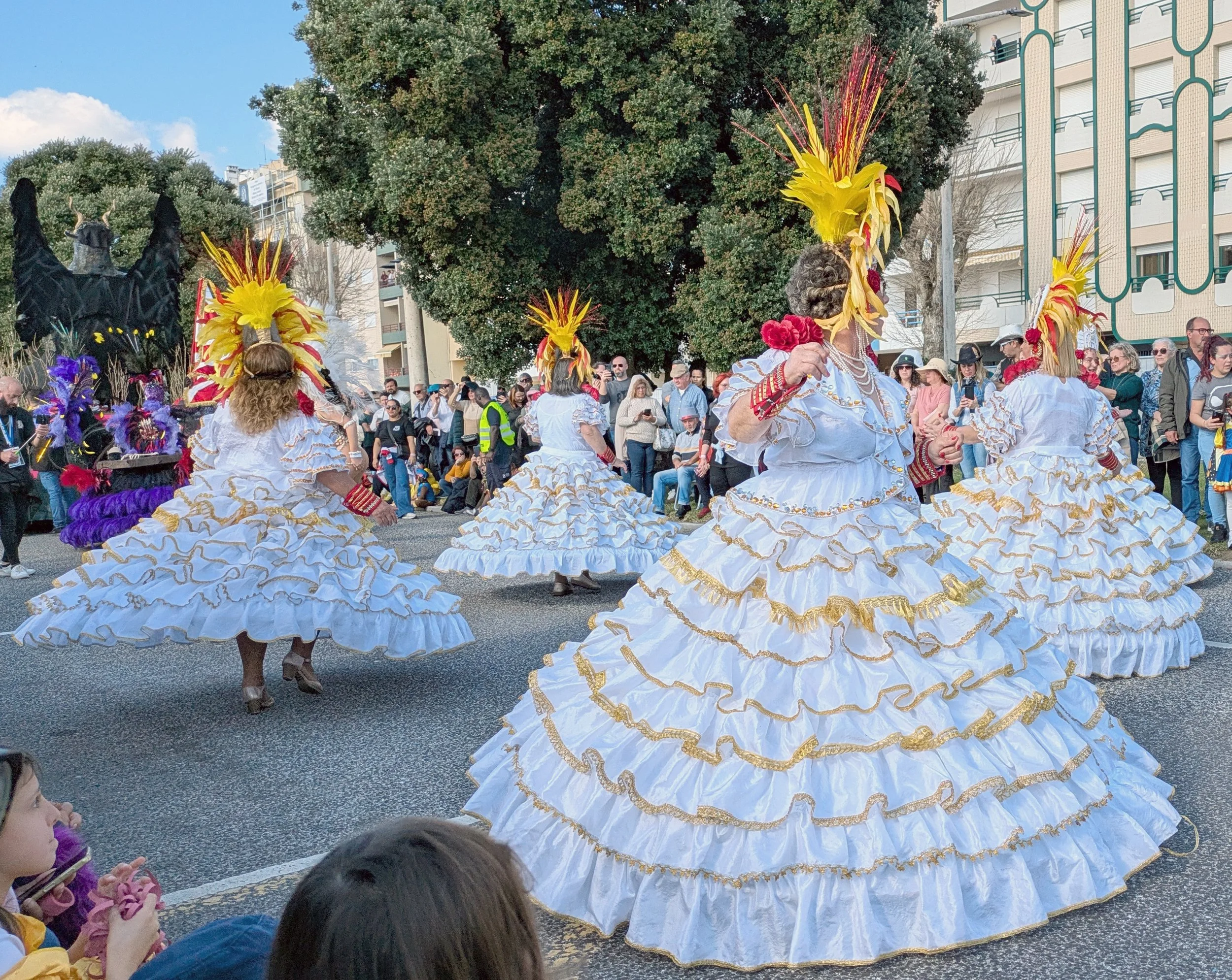 Women dressed in traditional white dresses with gold trim and large feathered yellow and red headdresses dance during a parade on a city street, with onlookers watching behind and buildings in the background.
