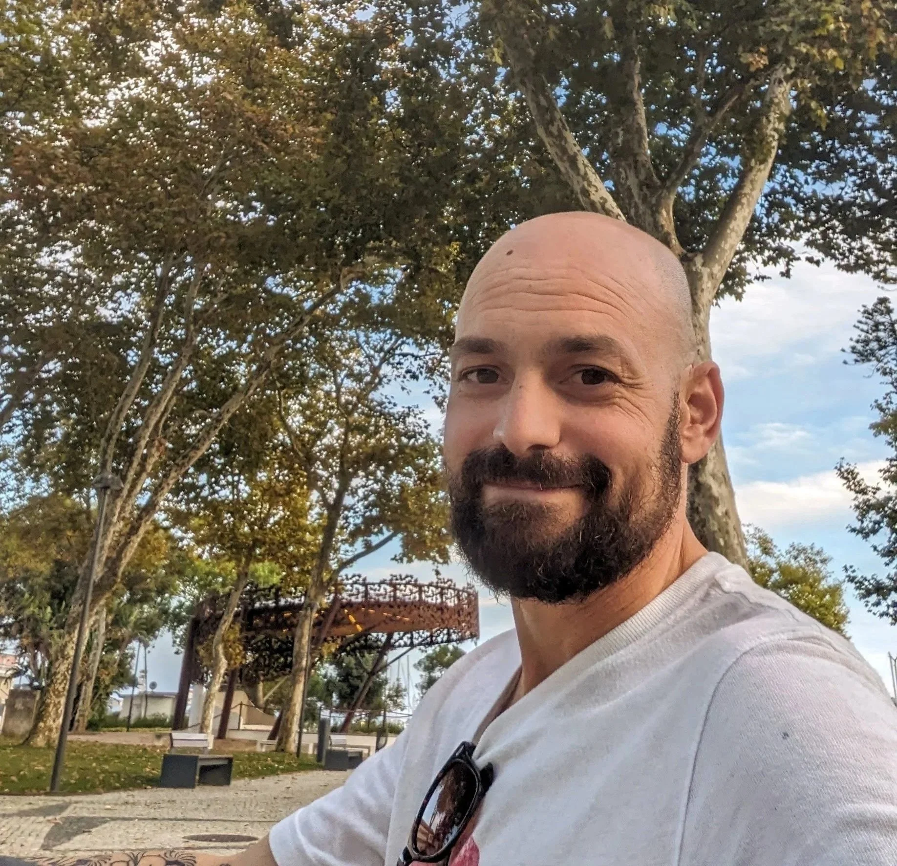 A man with a beard smiling outdoors in a park in Portugal with trees, benches, and a decorative metal structure in the background.