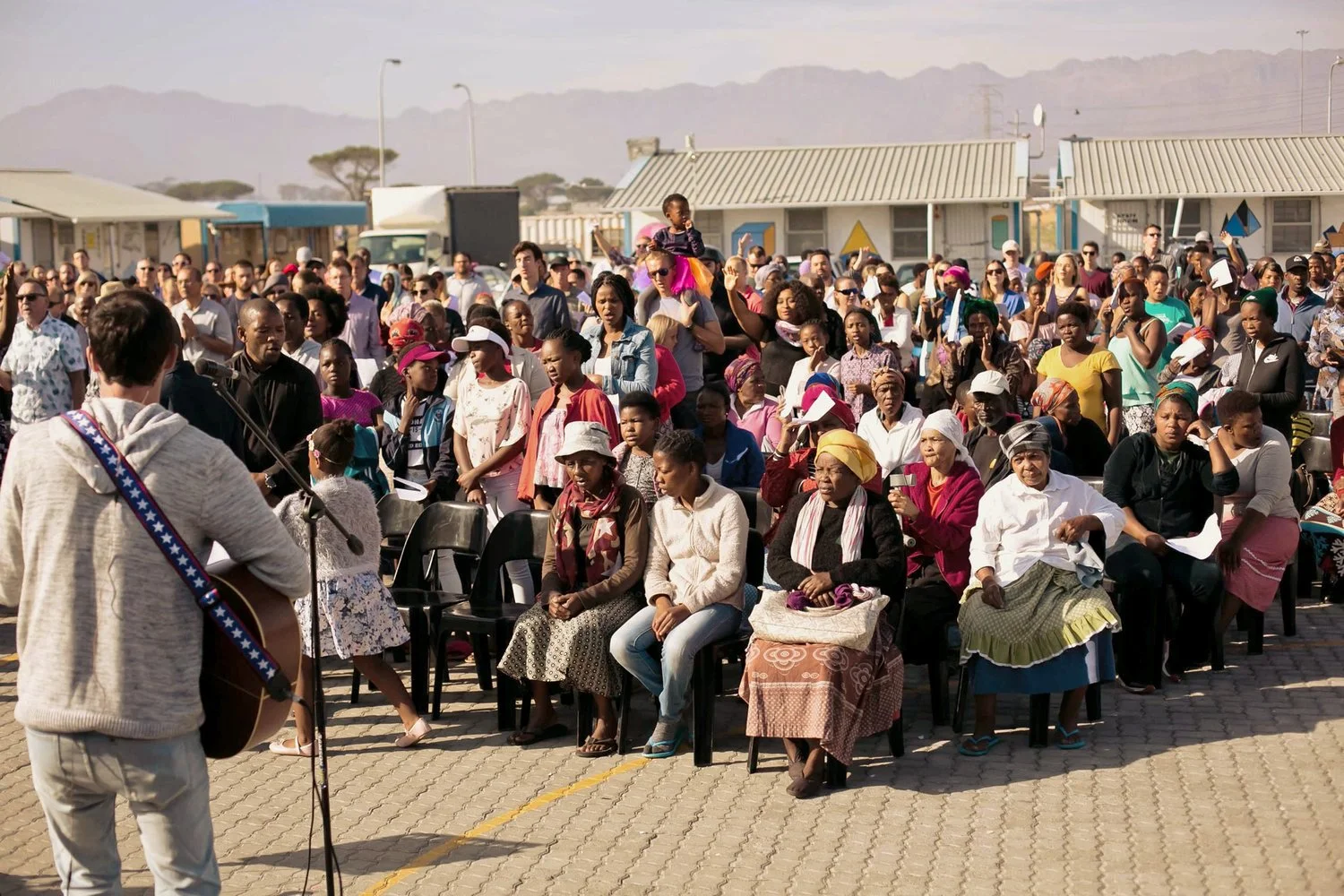 A large outdoor gathering with many people seated and standing, listening to a musician playing guitar and singing with a microphone, in a sunny setting with buildings and mountains in the background.
