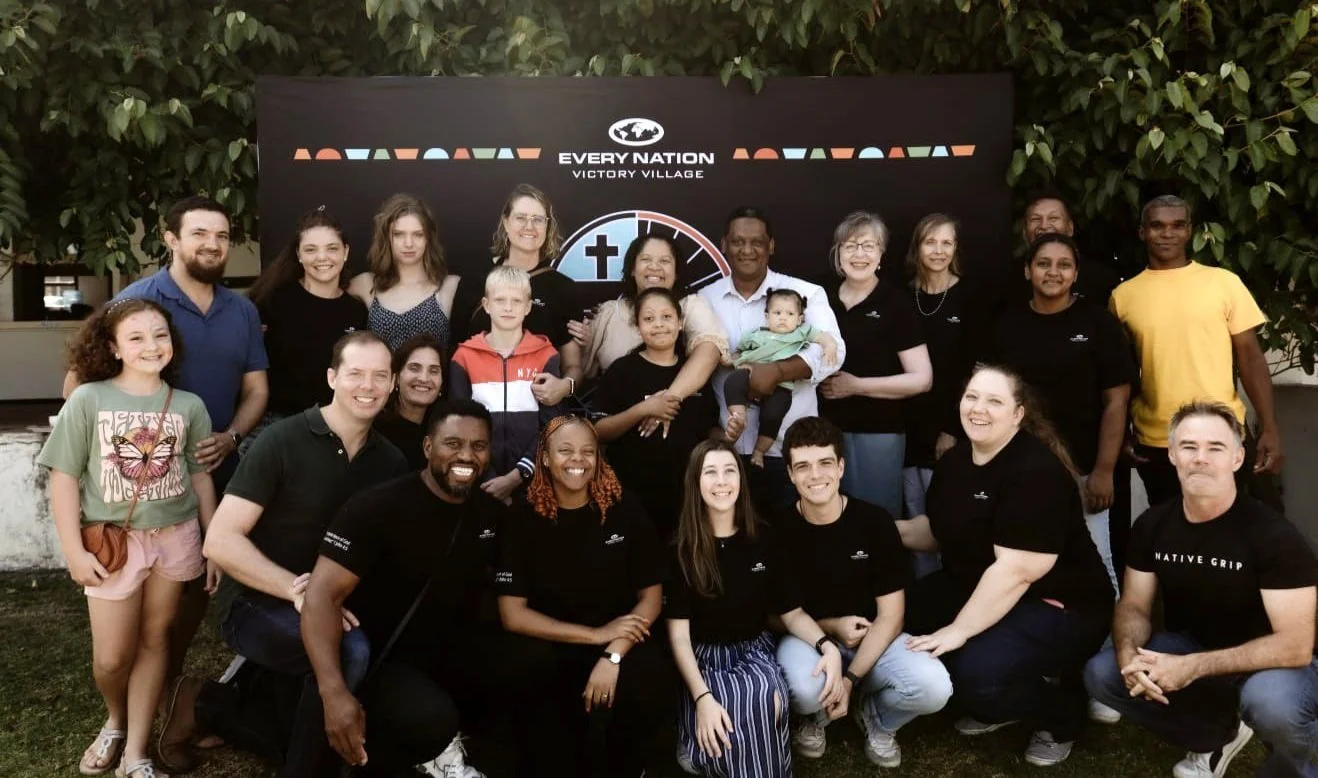 Group photo of diverse people, including children and adults, standing outdoors in front of a black backdrop with the words "Every Nation Victory Village" and a logo, smiling and posing for the camera.