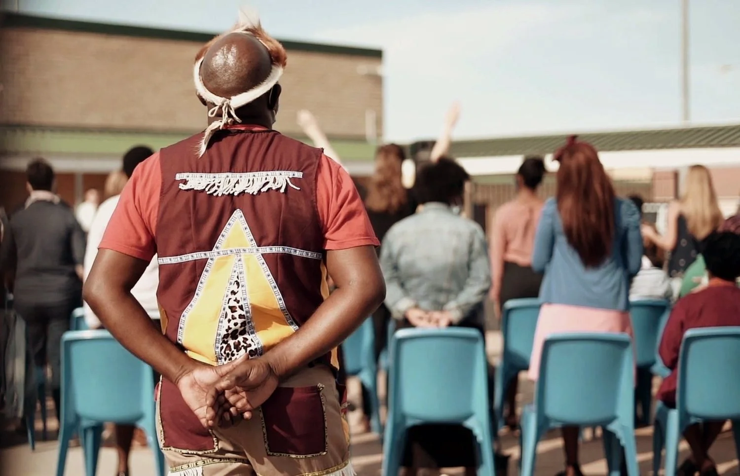 A person with a brown hairstyle and headband stands with their back to the camera and hands behind their back observing a group of people in an outdoor setting. The group of people involves men and women standing and sitting in front of a brick building, with some raising their hands, indicating a gathering or event.