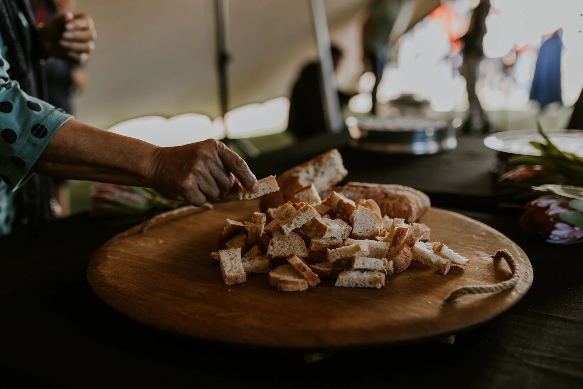 Person's hand cutting bread into pieces on a round wooden platter at a buffet or food station.