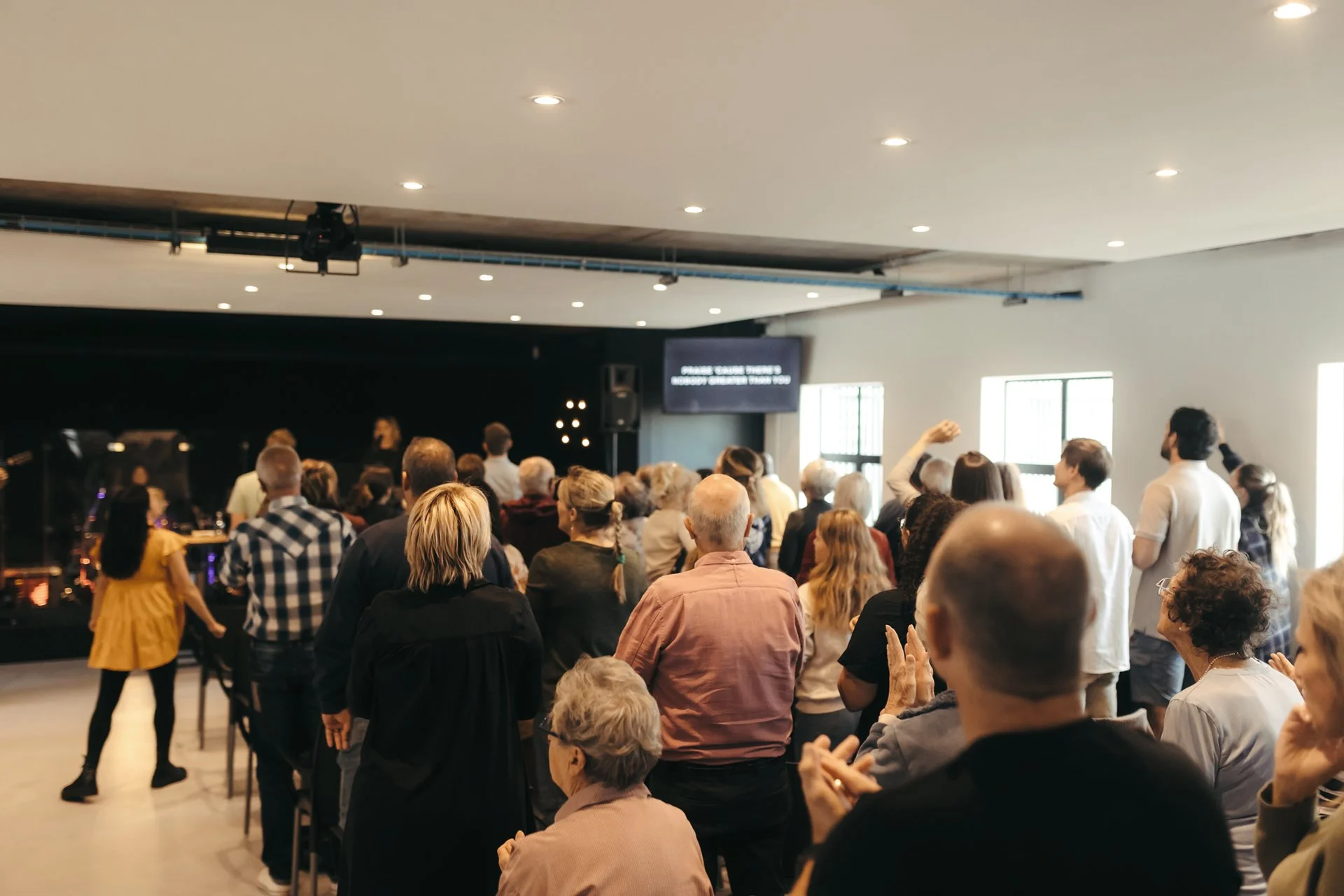 A group of people gathered in a room, watching a performance or presentation on stage. Some are standing, others are seated, and they are facing a dark stage area with a screen and microphones.