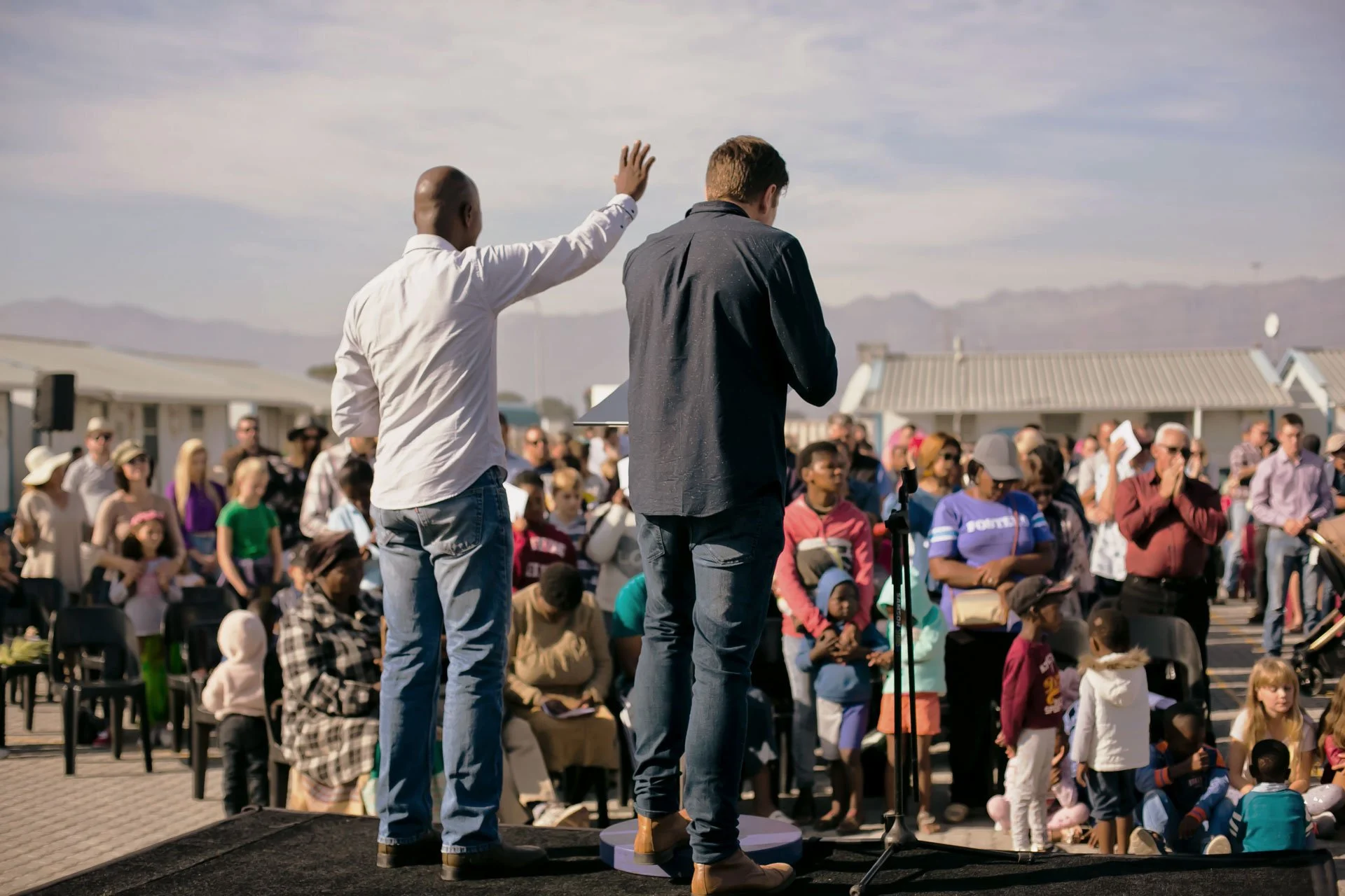 Two men are standing on a stage in front of a large crowd outdoors during daytime. One man is speaking and raising his hand, while the other is looking down. The audience includes children and adults, some seated and some standing, with houses and mountains visible in the background.