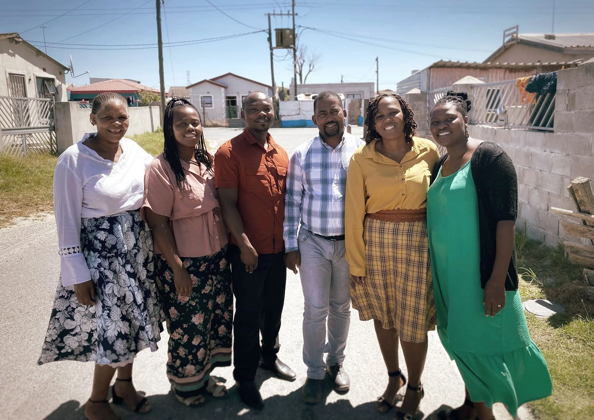 Group of seven diverse people standing outdoors on a sunny day, smiling and posing for a photo in a residential neighborhood.