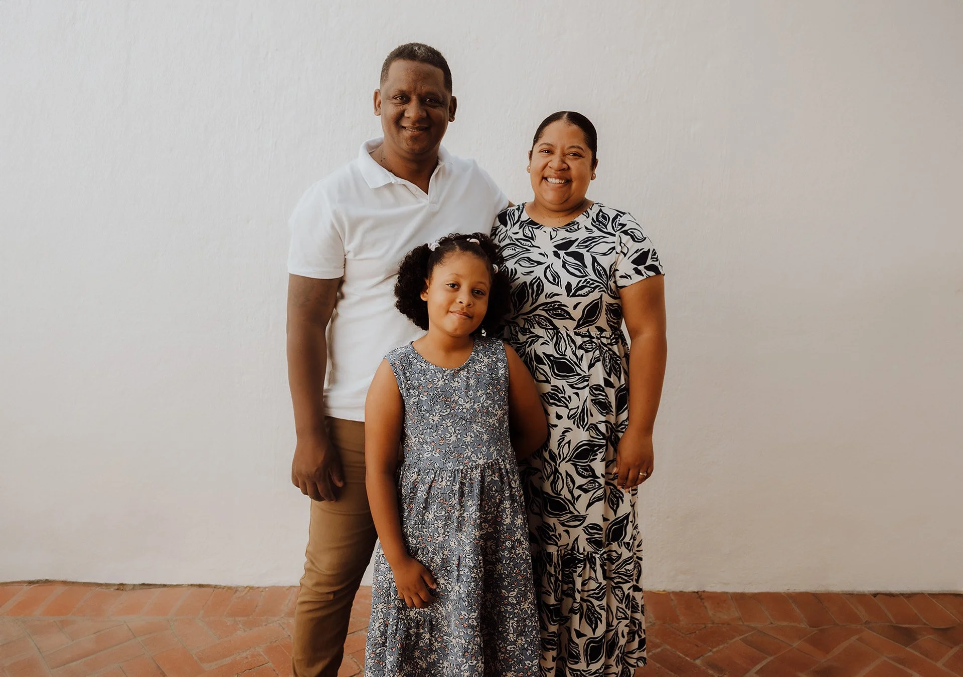 A family of three standing together, smiling, against a plain white wall. The man is on the left wearing a white polo shirt and khaki pants, the woman is on the right wearing a black and white patterned dress, and the young girl is in front wearing a blue floral dress.