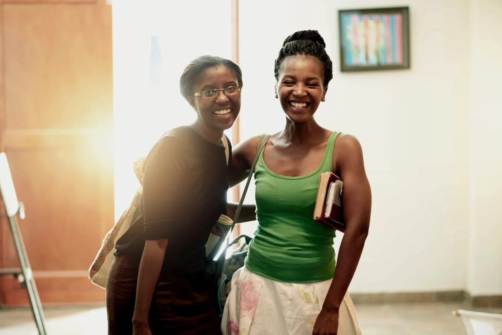 Two women smiling and standing together in an indoor setting, one wearing glasses and the other in a green tank top, both holding notebooks and backpacks.