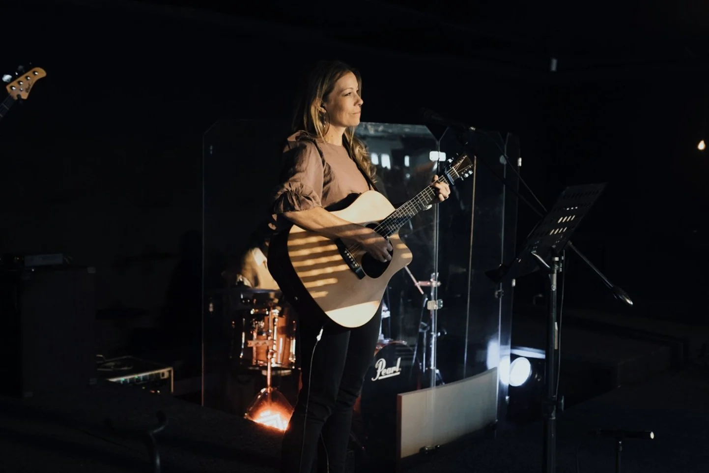 A woman playing an acoustic guitar on a dimly lit stage, with a drum set and music stand nearby, and a clear protective screen around her.