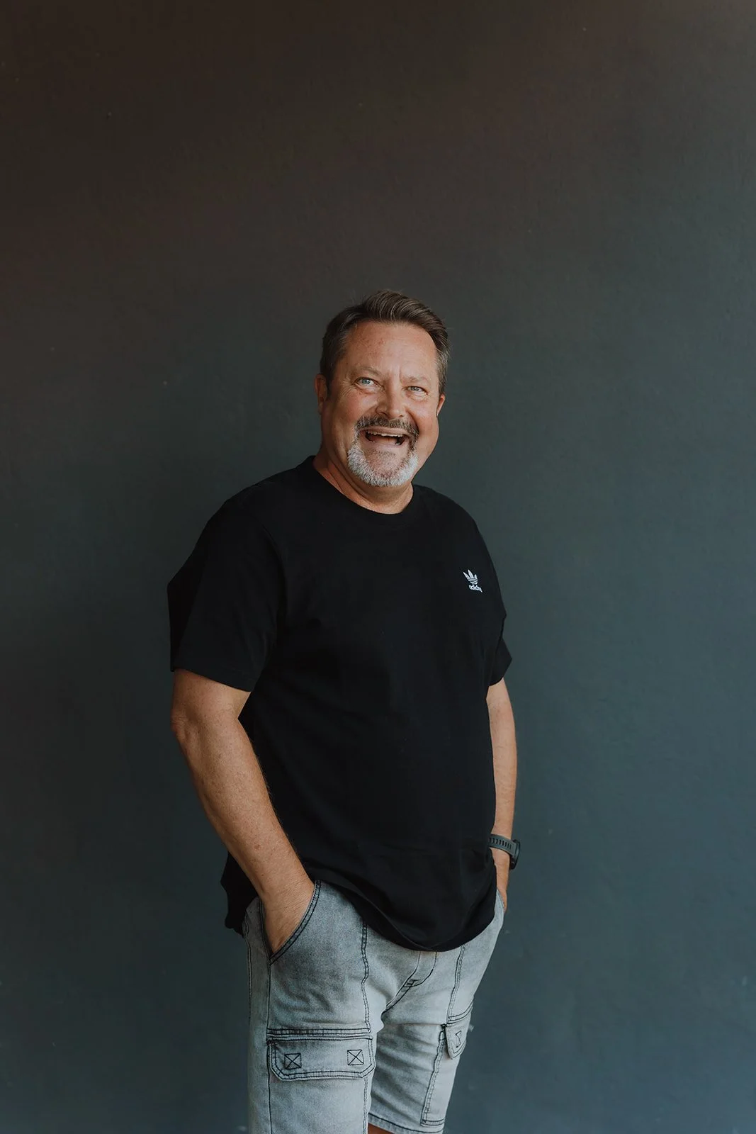 A family of three standing together, smiling, against a plain white wall. The man is on the left wearing a white polo shirt and khaki pants, the woman is on the right wearing a black and white patterned dress, and the young girl is in front wearing a blue floral dress.