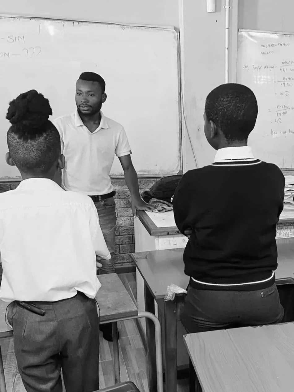 A classroom scene with a teacher standing and explaining in front of three students who are paying attention. There are whiteboards with writing behind him.