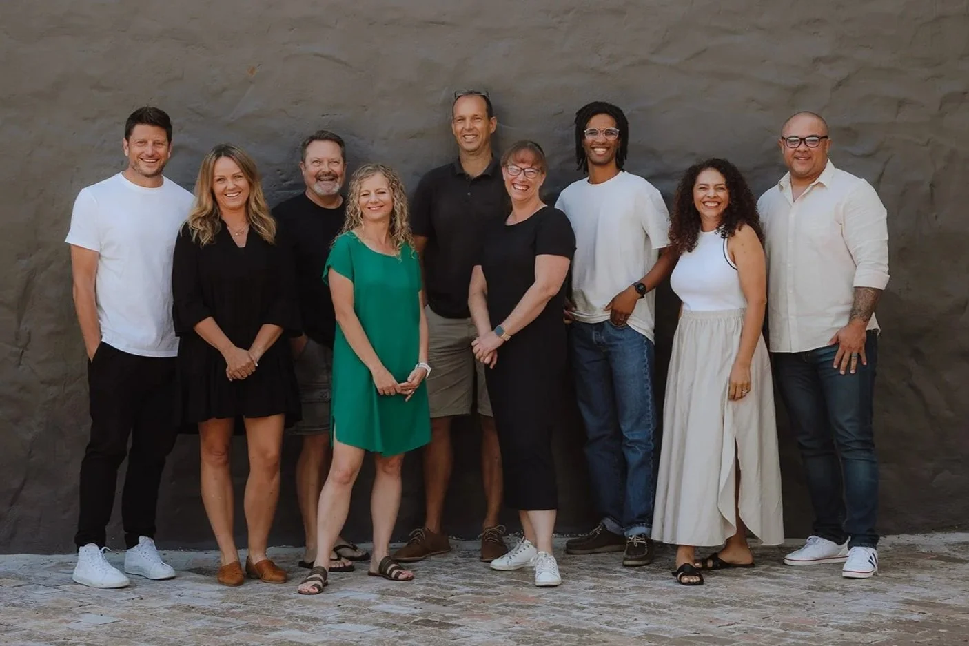 Group of ten diverse people standing together outdoors against a stone wall, smiling at the camera.