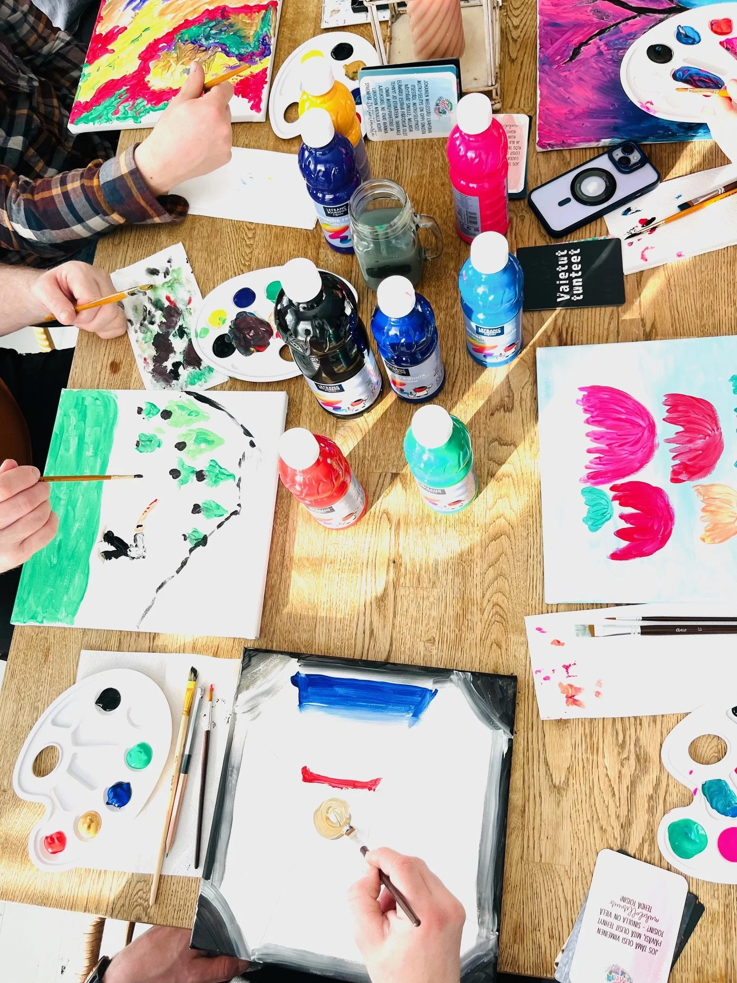 People painting bright, colorful artworks on paper with paint palettes, brushes, and bottles of paint on a wooden table.