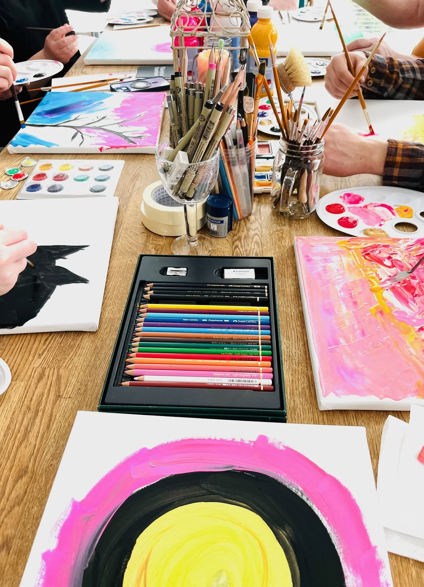 A group of people painting abstract art on canvases with colorful paints and brushes on a wooden table, surrounded by art supplies like colored pencils, paints, and markers.