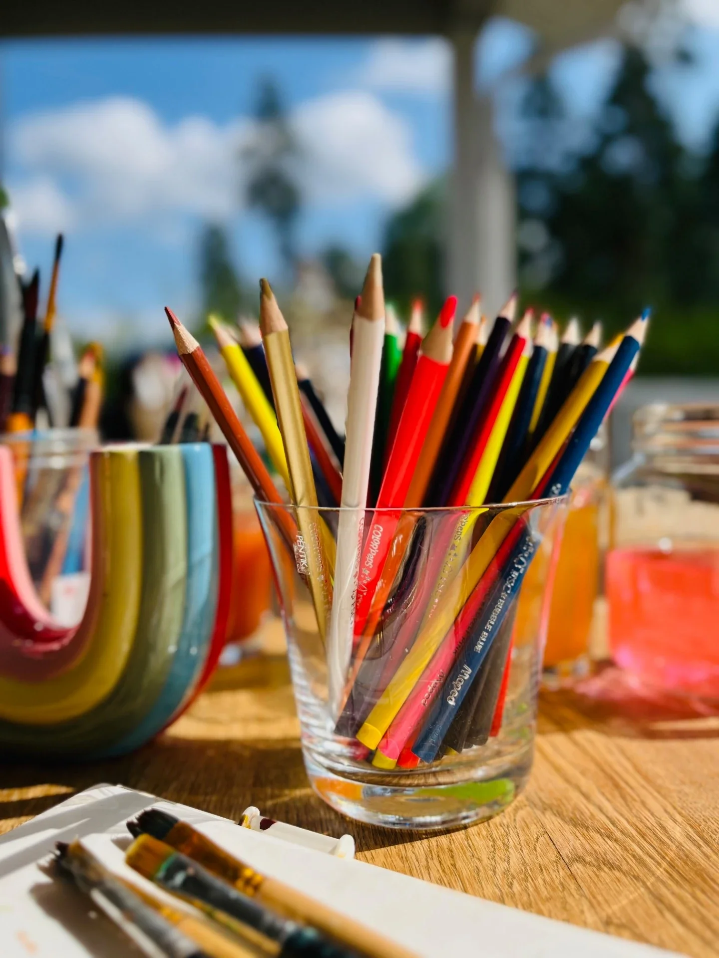 A glass filled with colorful pencils on a wooden table outdoors with a blue sky and clouds in the background.