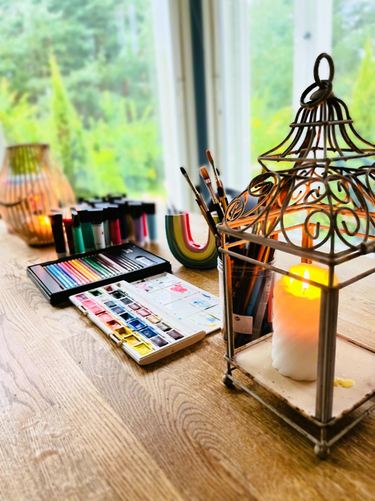 Art supplies on a wooden table, including watercolor paints, brushes, and colored paper, with a window view of greenery outside.