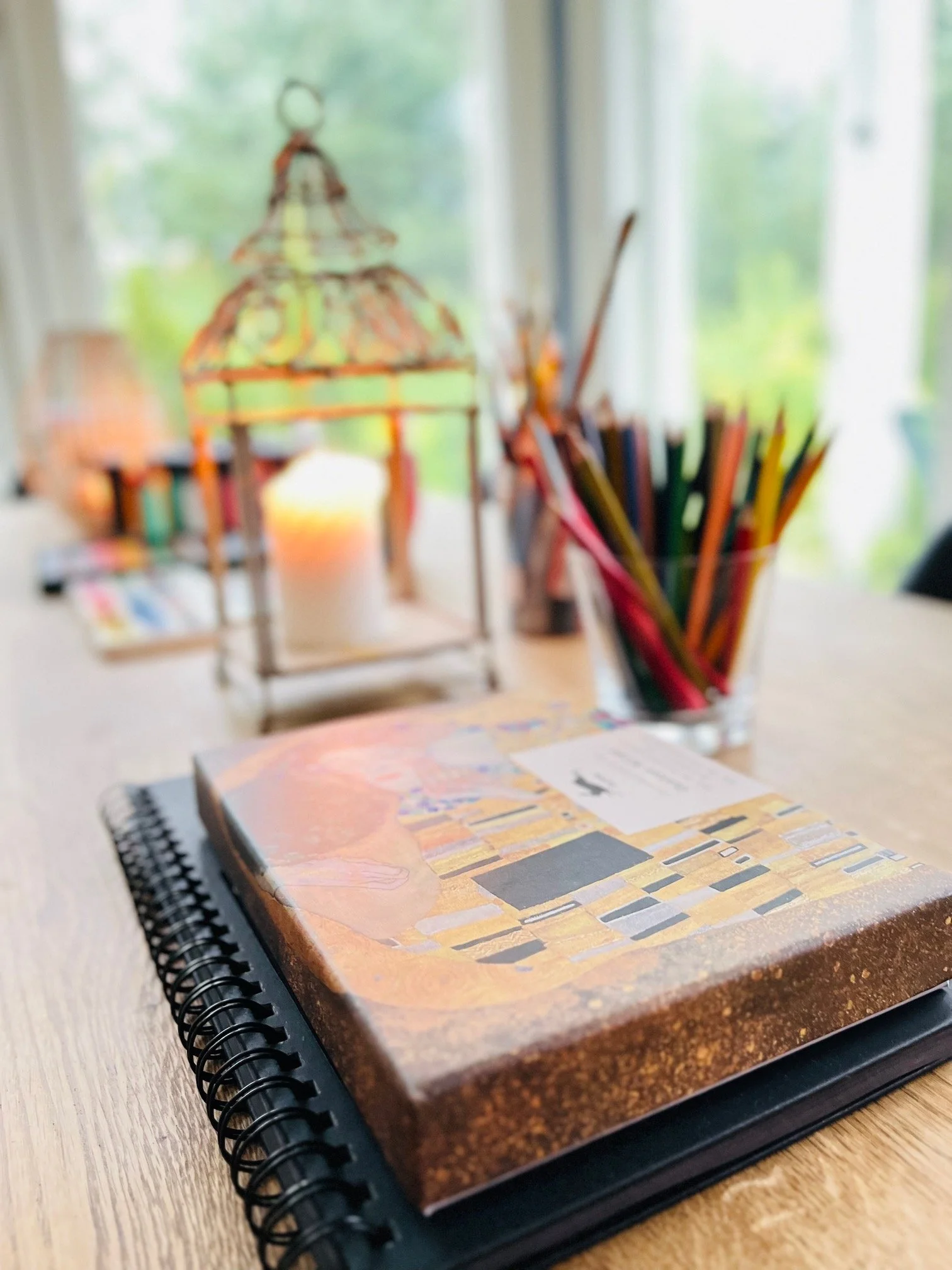 A spiral-bound art journal with artwork on the cover placed on a wooden table, with a cup of colored pencils and a decorative lantern with a candle inside on a bright windowsill.