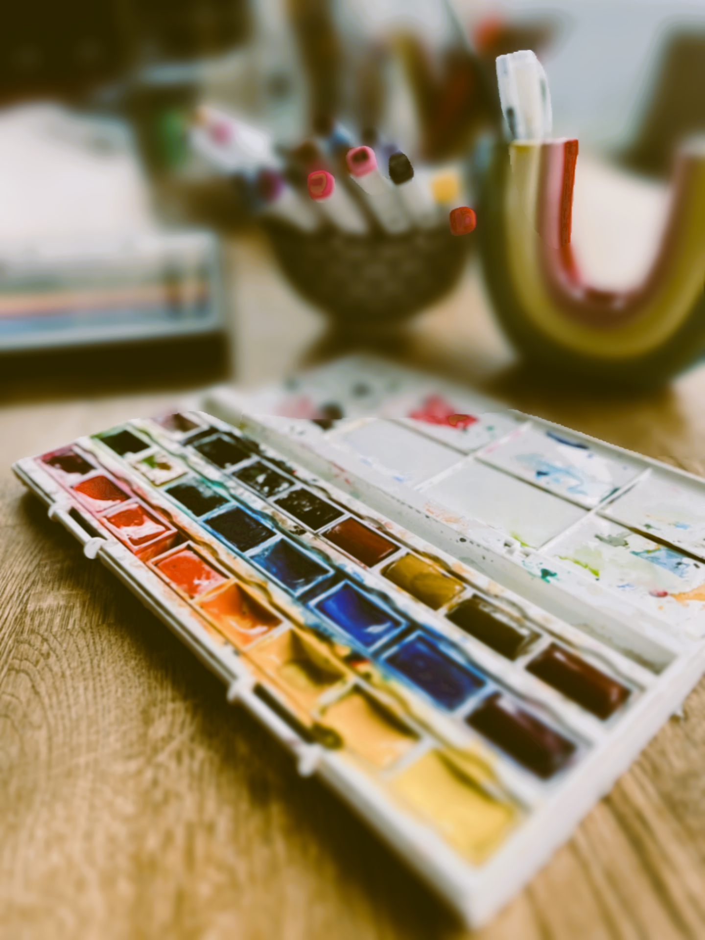 A watercolor paint set with various colors, a holder with markers or colored pens, a cup or container, and a decorative gold-colored letter 'U' on a wooden surface.