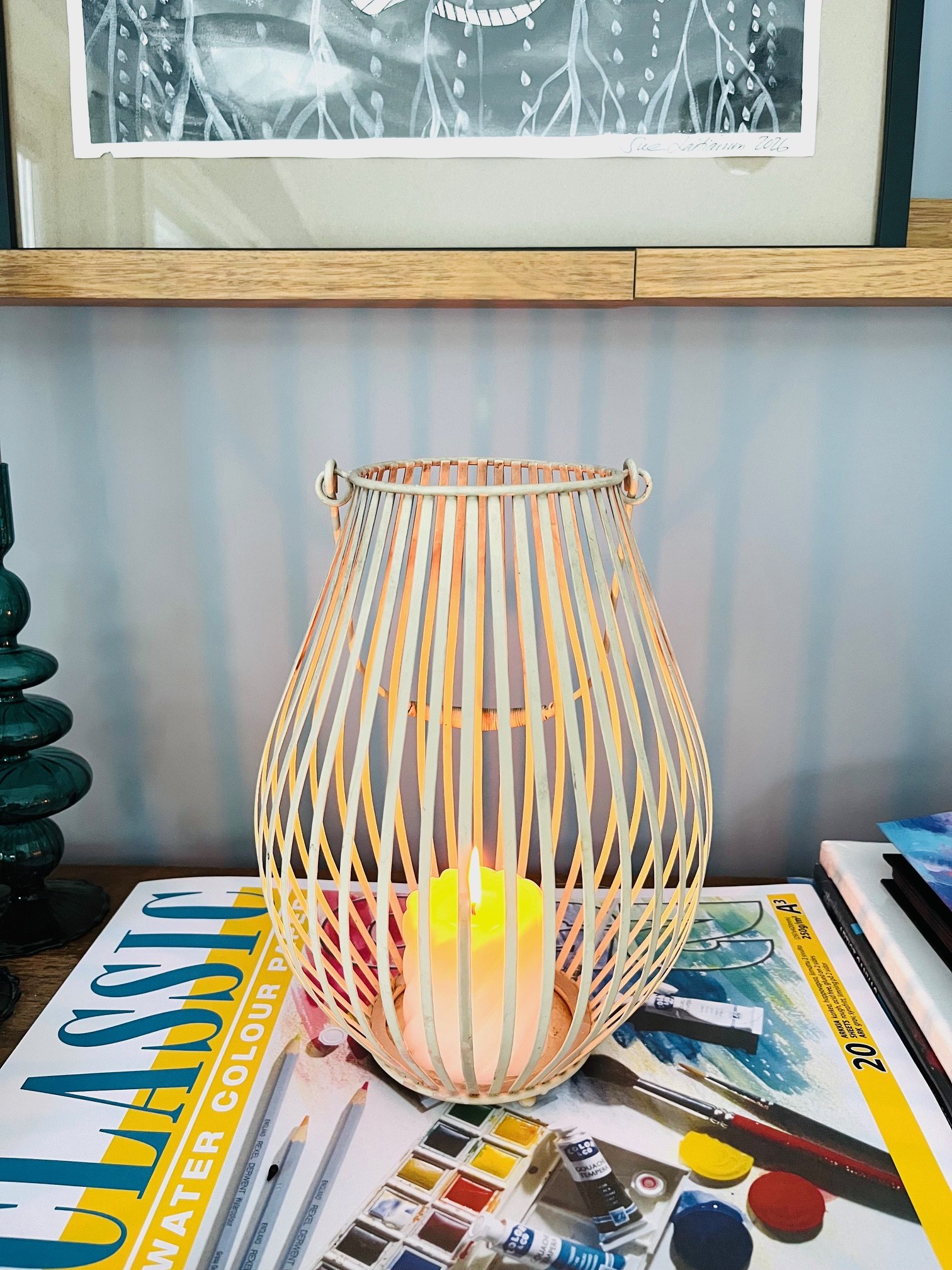 Decorative lamp with a candle inside, placed on a table covered with a spread about watercolors and art supplies, with a framed artwork hanging on the wall above.